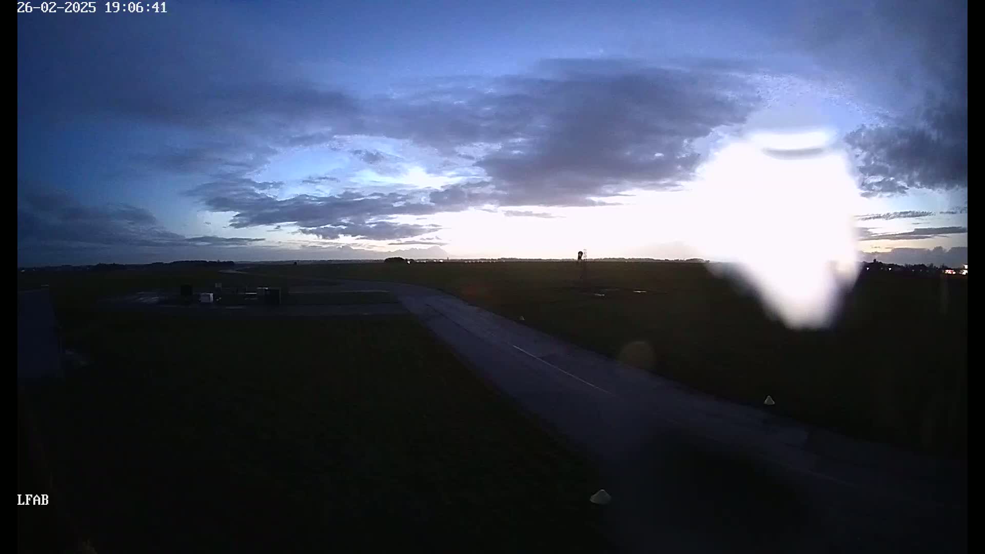A dark, flat landscape with a road and a bright, blurry area in the sky under a twilight sky with mostly cloudy conditions.