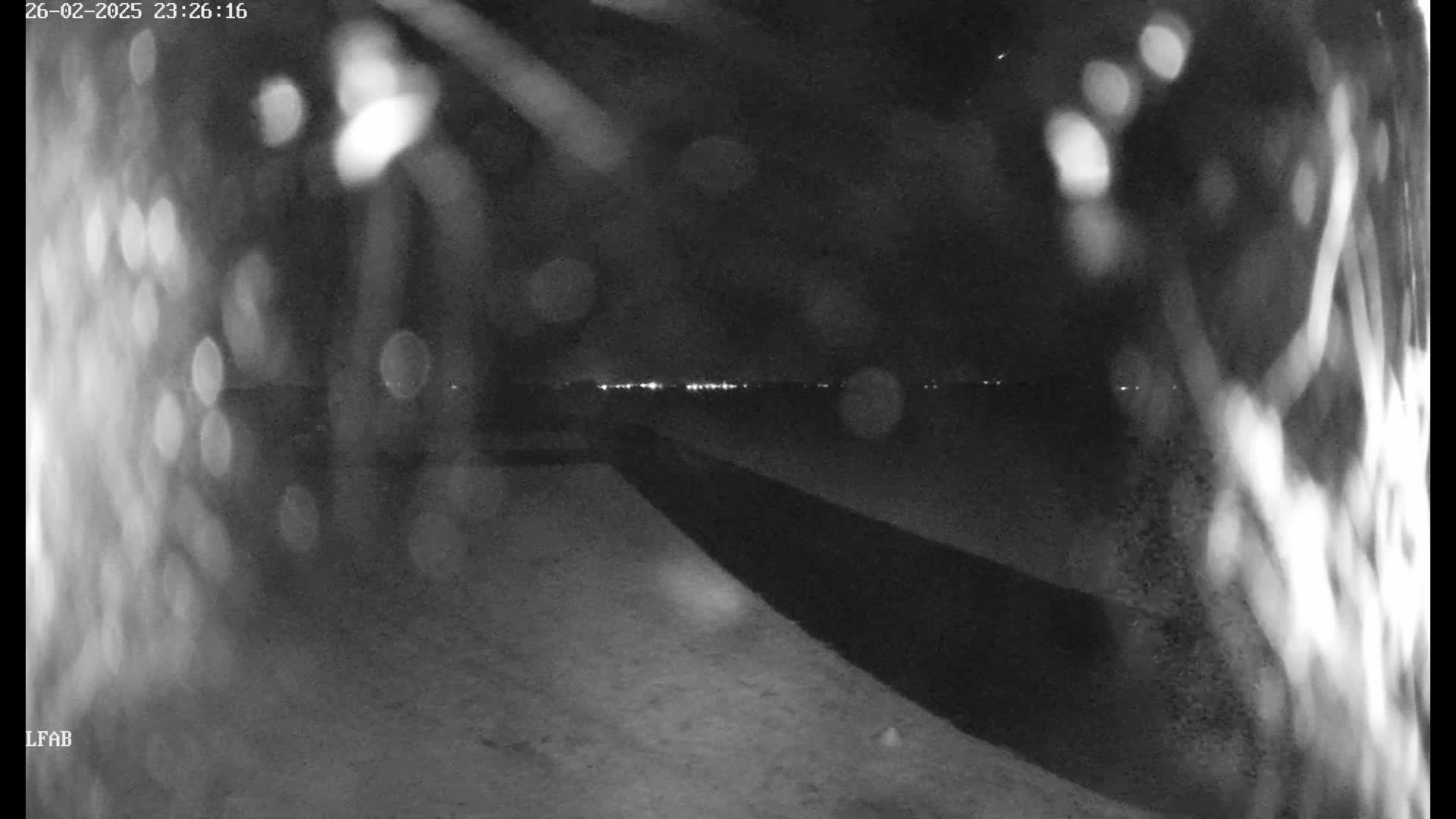 A nighttime, blurry view shows a dark, possibly snow-covered ground leading toward a distant line of lights under a dark sky with some rain or snow obscuring the view.