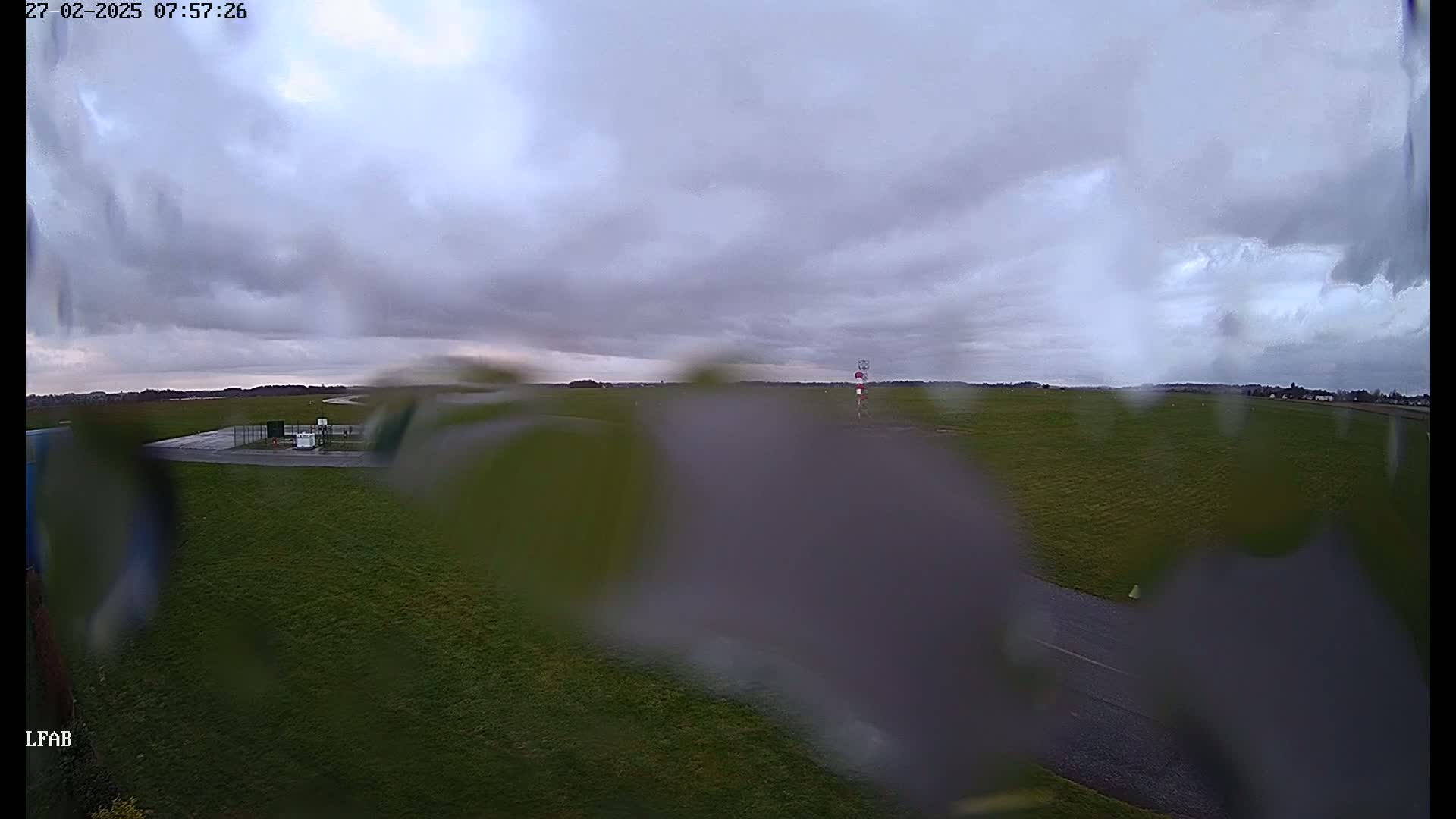 A rain-covered lens obscures the view of a grassy airfield under an overcast sky.