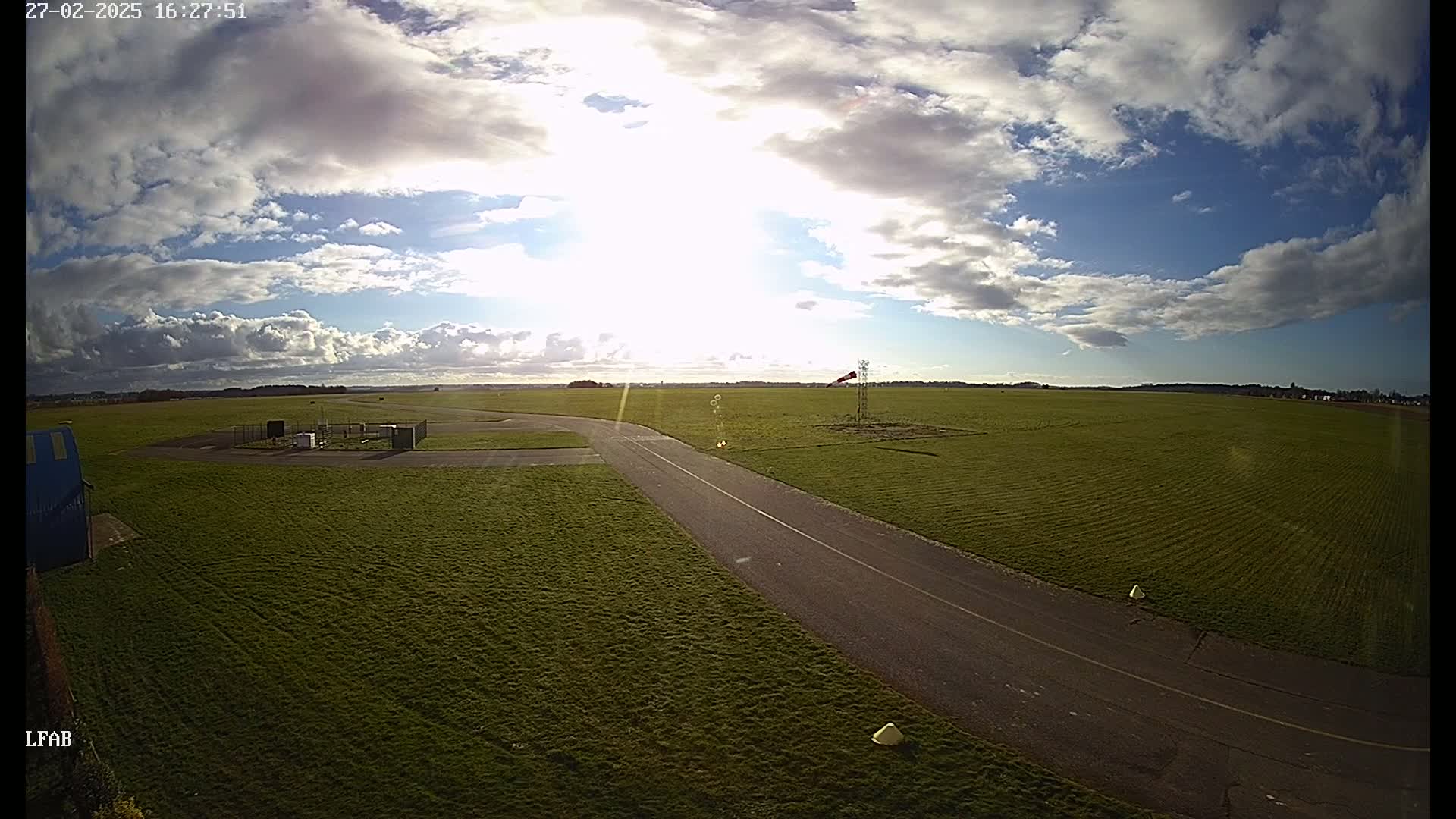 A paved road curves through a grassy airfield under a partly sunny sky with scattered clouds.