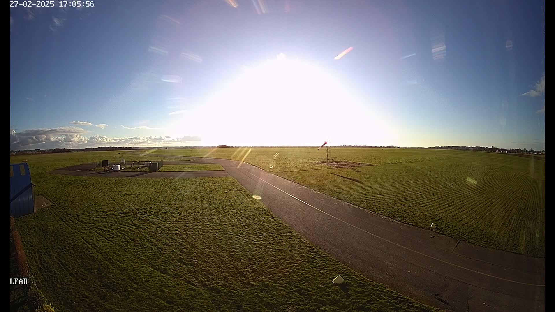 A sunny day at a grassy airfield shows a paved taxiway curving through a field with a windsock visible in the distance.