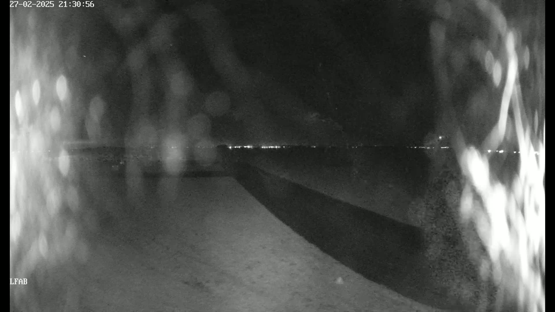 A nighttime, blurry view shows a dark, possibly paved area leading to a distant horizon where faint lights are visible, with what appears to be rain or moisture obscuring the view.