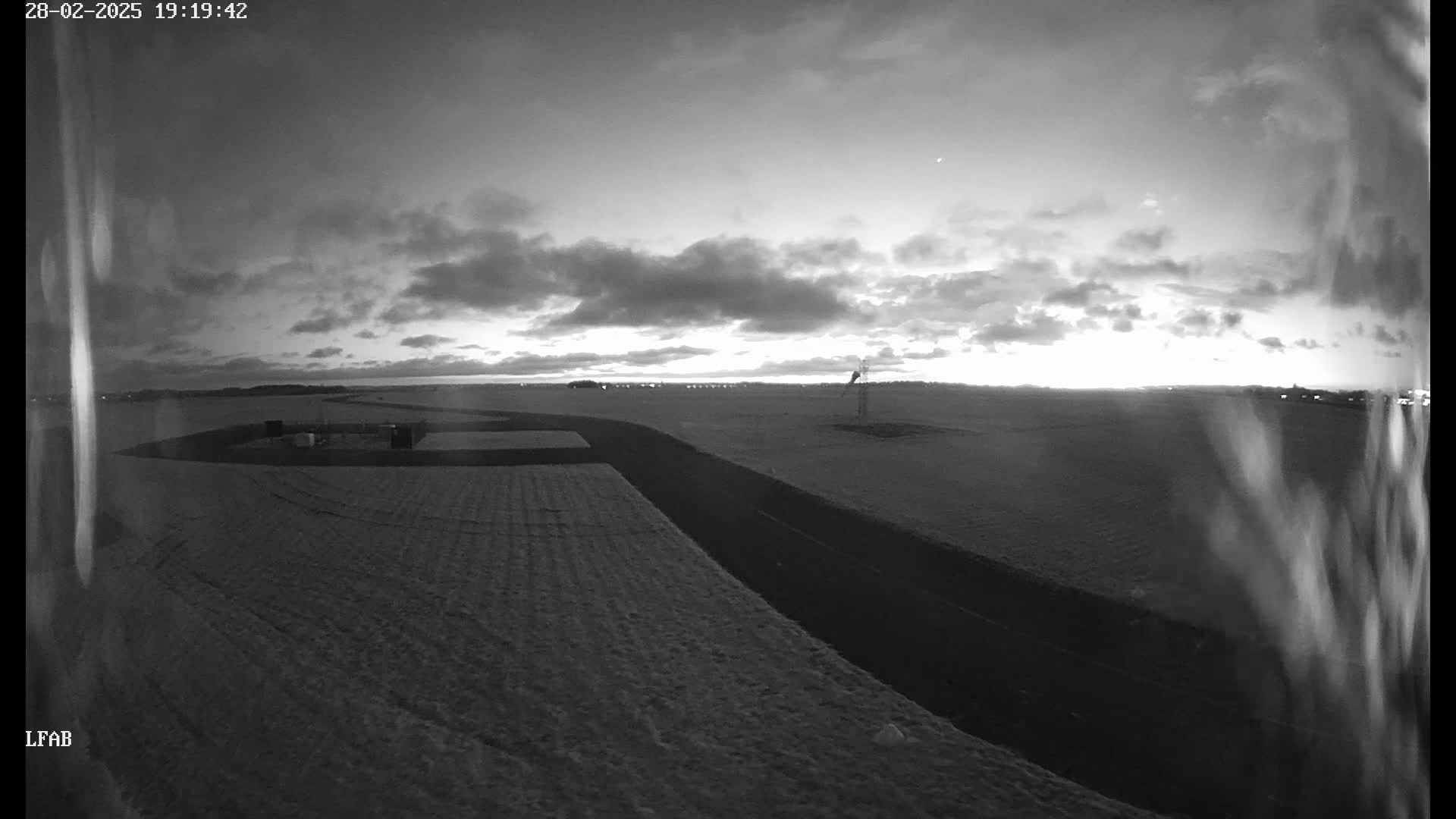 A black and white, nighttime image shows a flat, snow-covered landscape with a curving road and a distant structure under a cloudy sky.