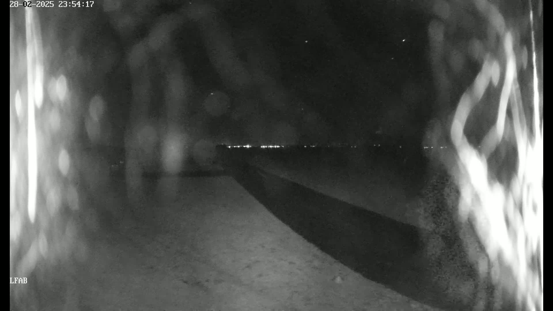 A nighttime, low-light image shows a dark, paved area extending into the distance towards a faintly visible line of lights under a clear, dark sky.