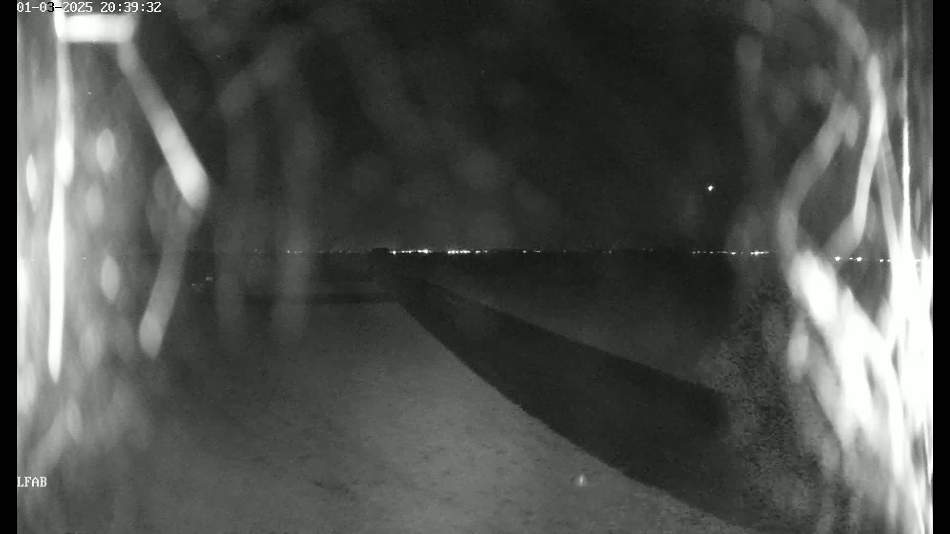 A nighttime, low-light view shows a dark, flat expanse of land with a long, dark shadow cast across it, leading toward a distant horizon illuminated by faint lights.
