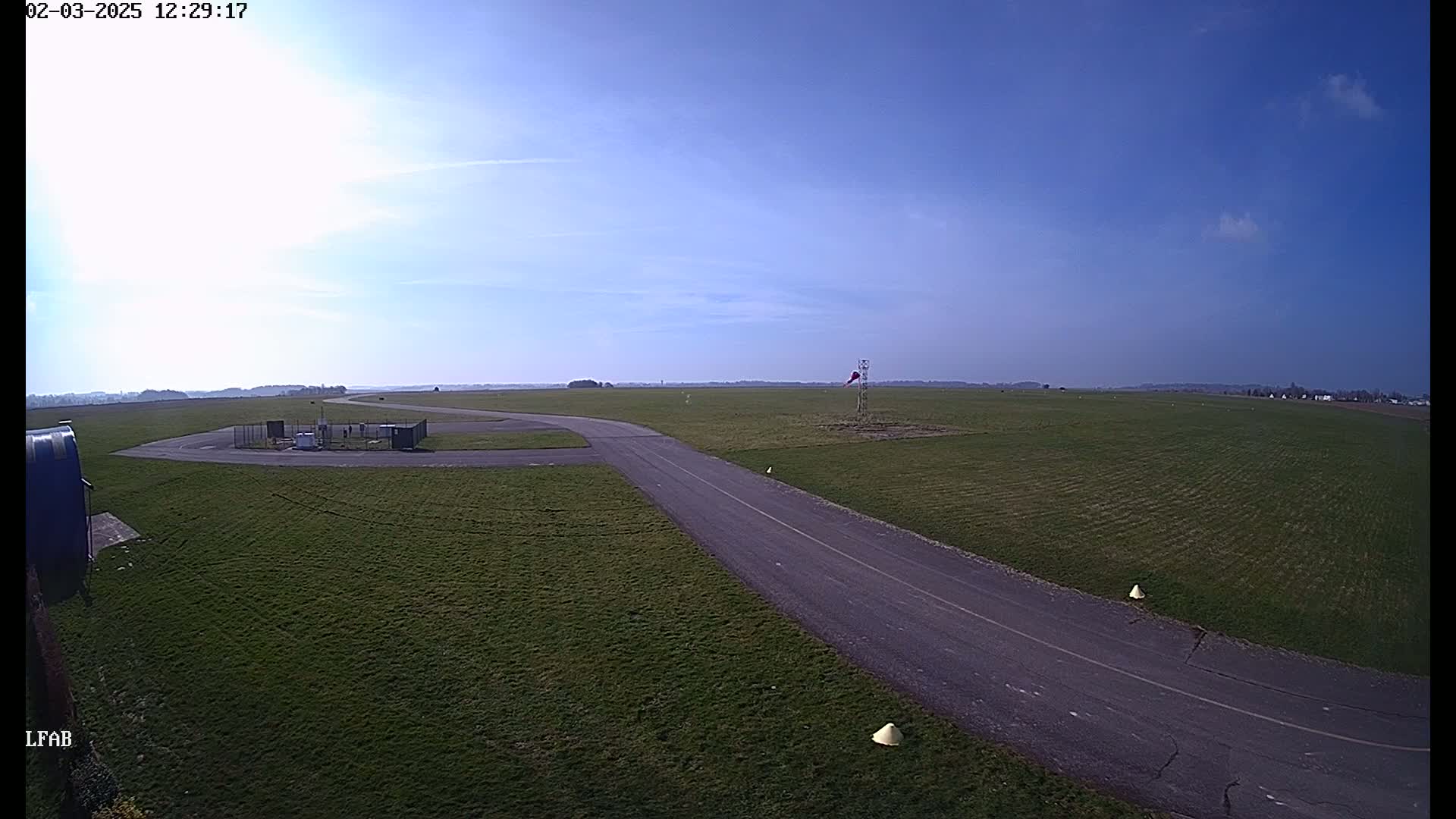 A paved taxiway curves through a grassy airfield under a mostly clear, bright blue sky.