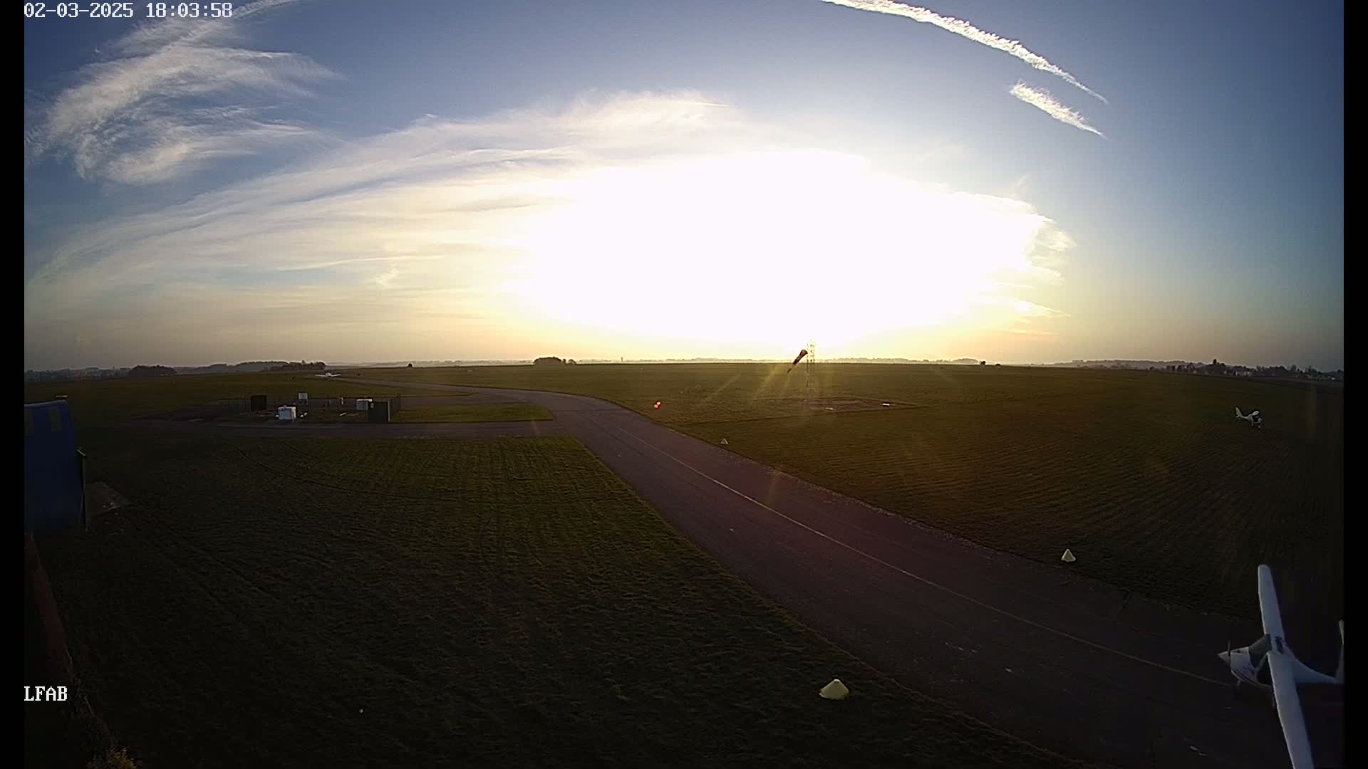A high-angle, wide shot shows a grassy airfield at sunset under partly cloudy skies, with two small airplanes on the ground and a taxiway leading toward the sun.