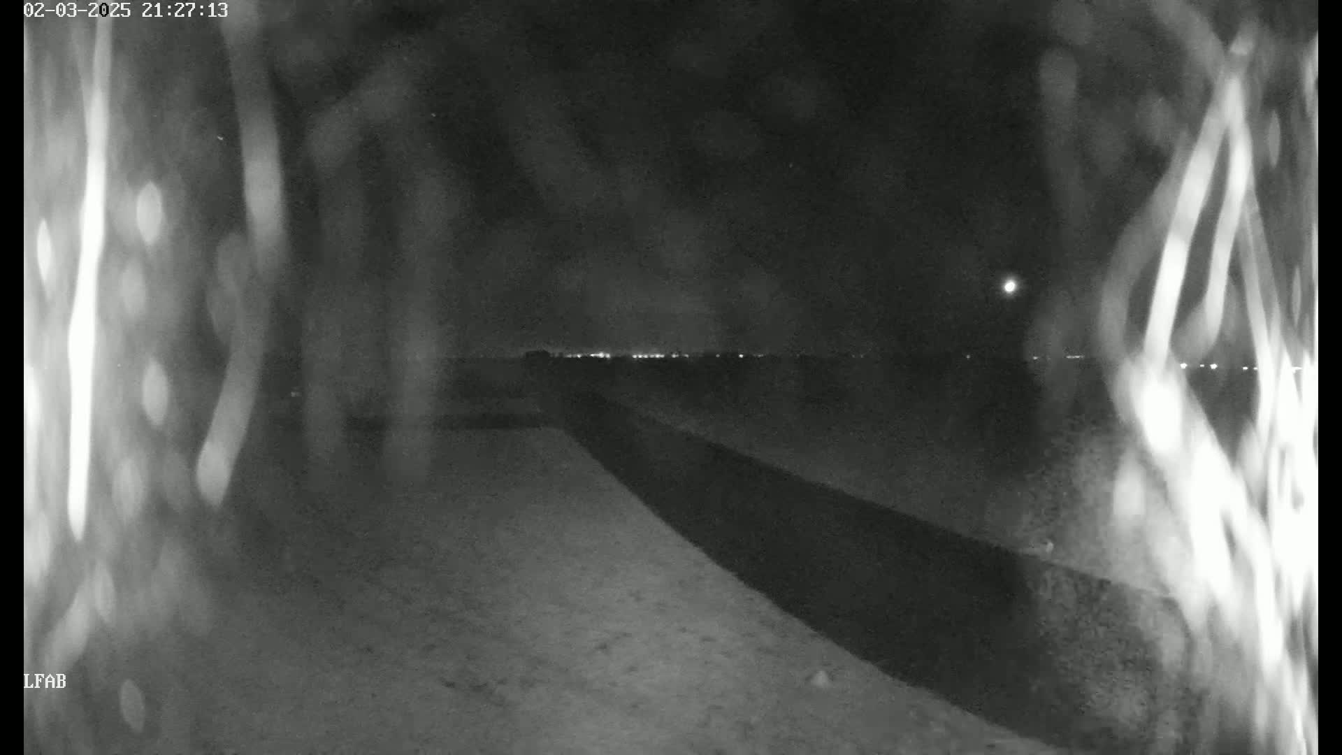 A nighttime, low-light view shows a dark, possibly paved, area extending into the distance, with a line of distant lights visible on the horizon.