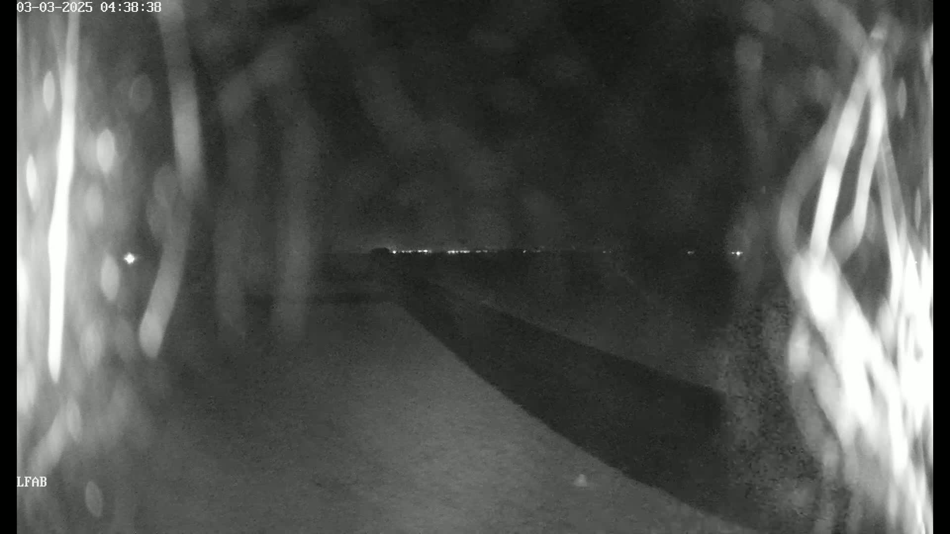 A nighttime, low-light view shows a dark road or path extending to a distant line of lights, partially obscured by blurred, indistinct foreground objects.