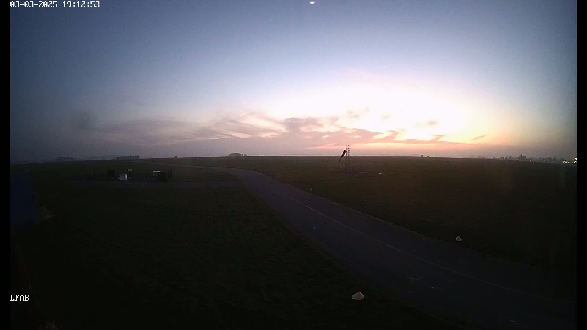 A long, dark road extends from the foreground into a flat, grassy field under a twilight sky with a few clouds and a hint of pink near the horizon.
