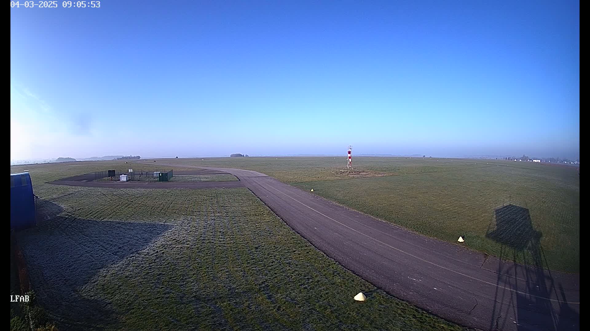 A frost-covered airfield with a paved taxiway stretches toward a distant horizon under a clear blue sky.
