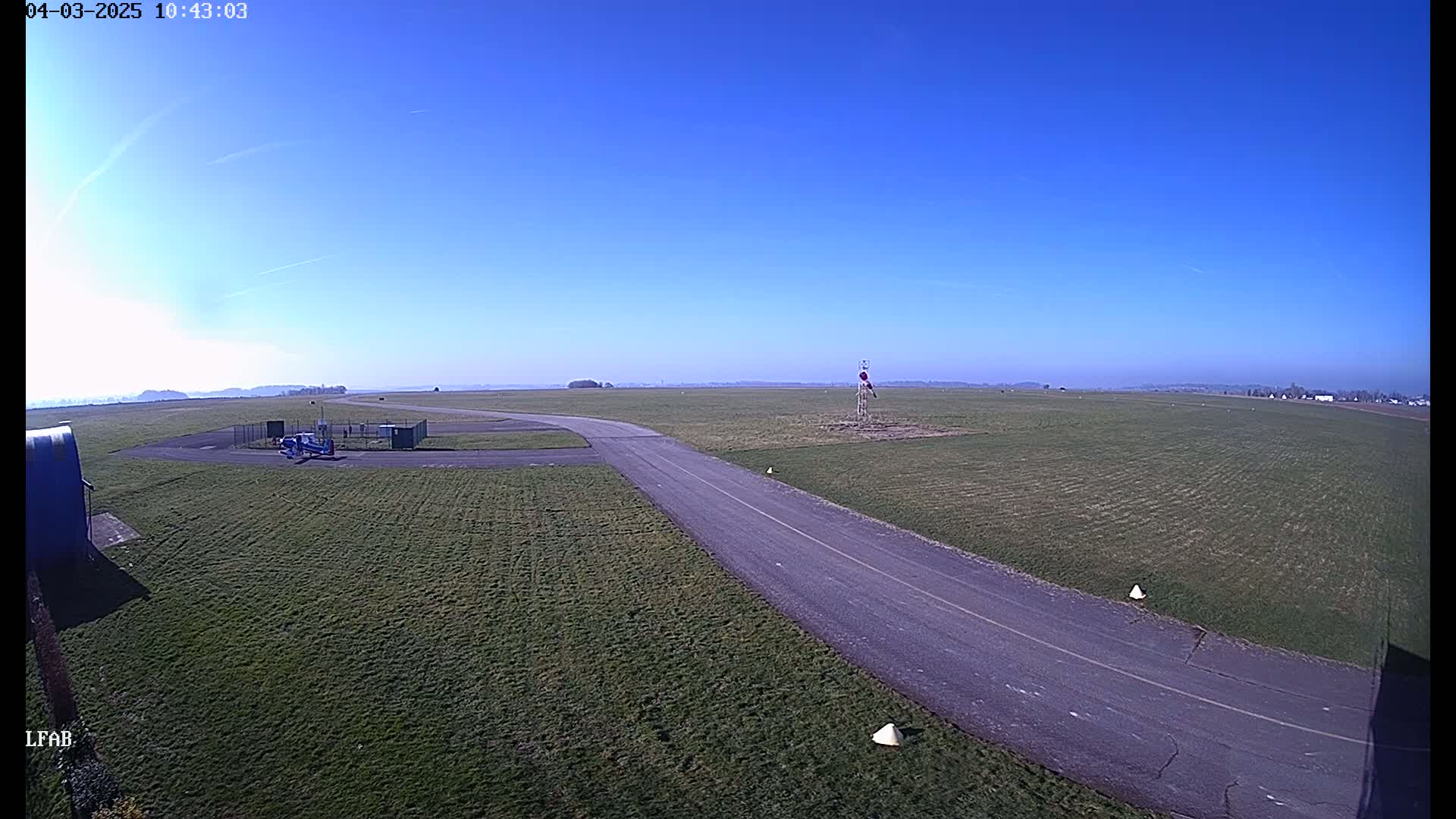 A small airplane sits on a grassy airfield on a clear, sunny day, with a paved taxiway curving towards the horizon.