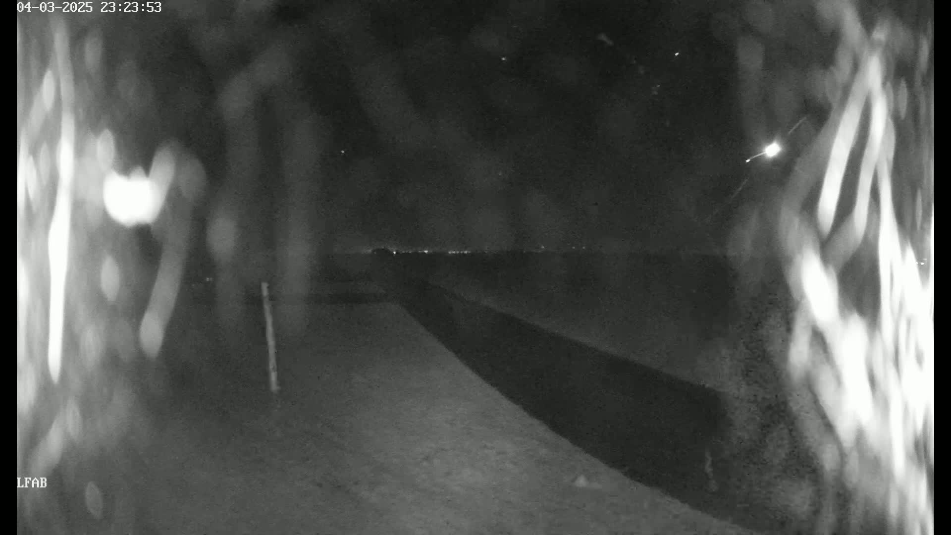 A nighttime, low-light view shows a dark road leading to a distant horizon of lights under a clear sky with a few visible stars and a streak of light possibly from a meteor.