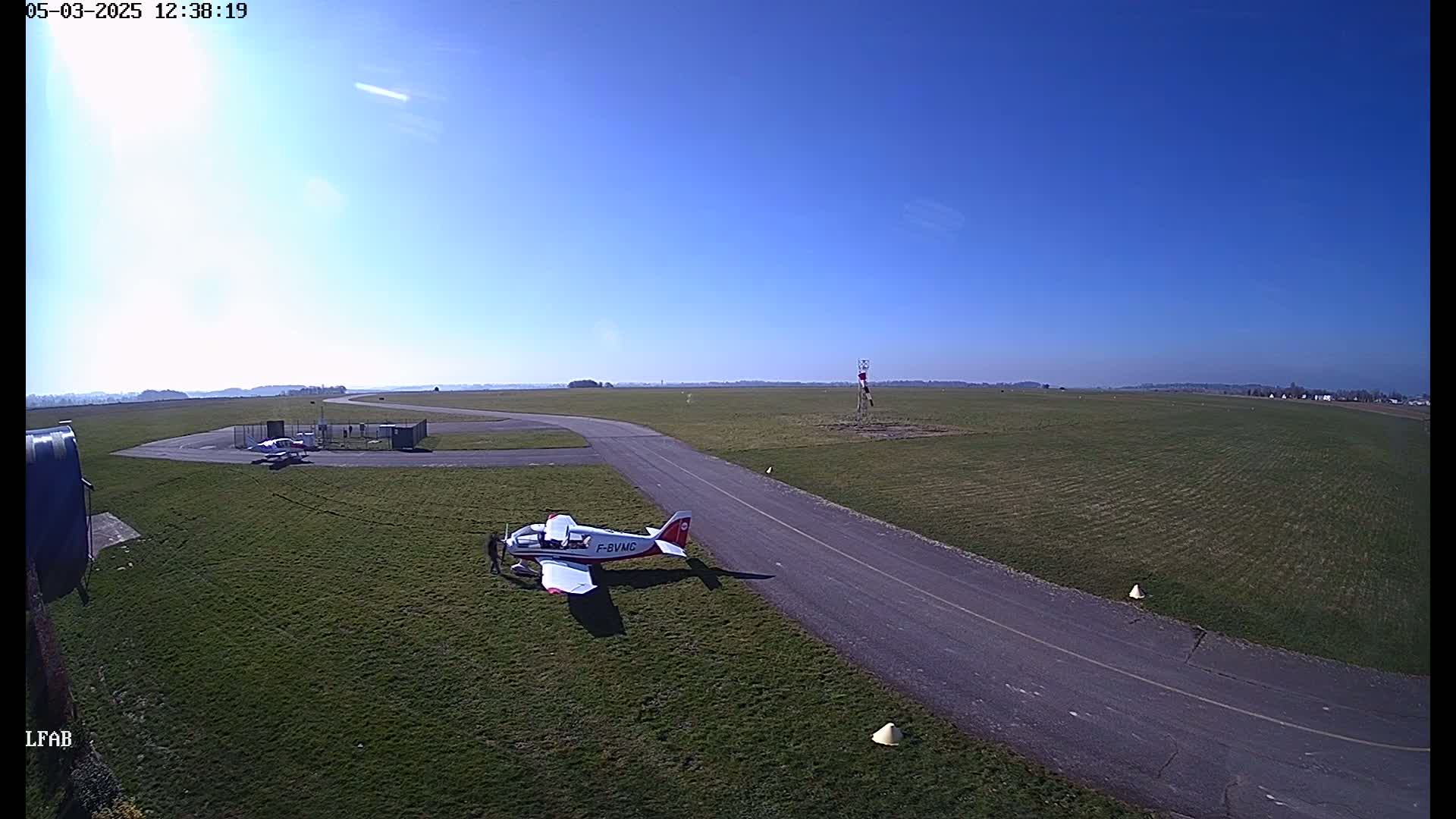 A small airplane sits on a grassy airfield taxiway under a clear blue sky.
