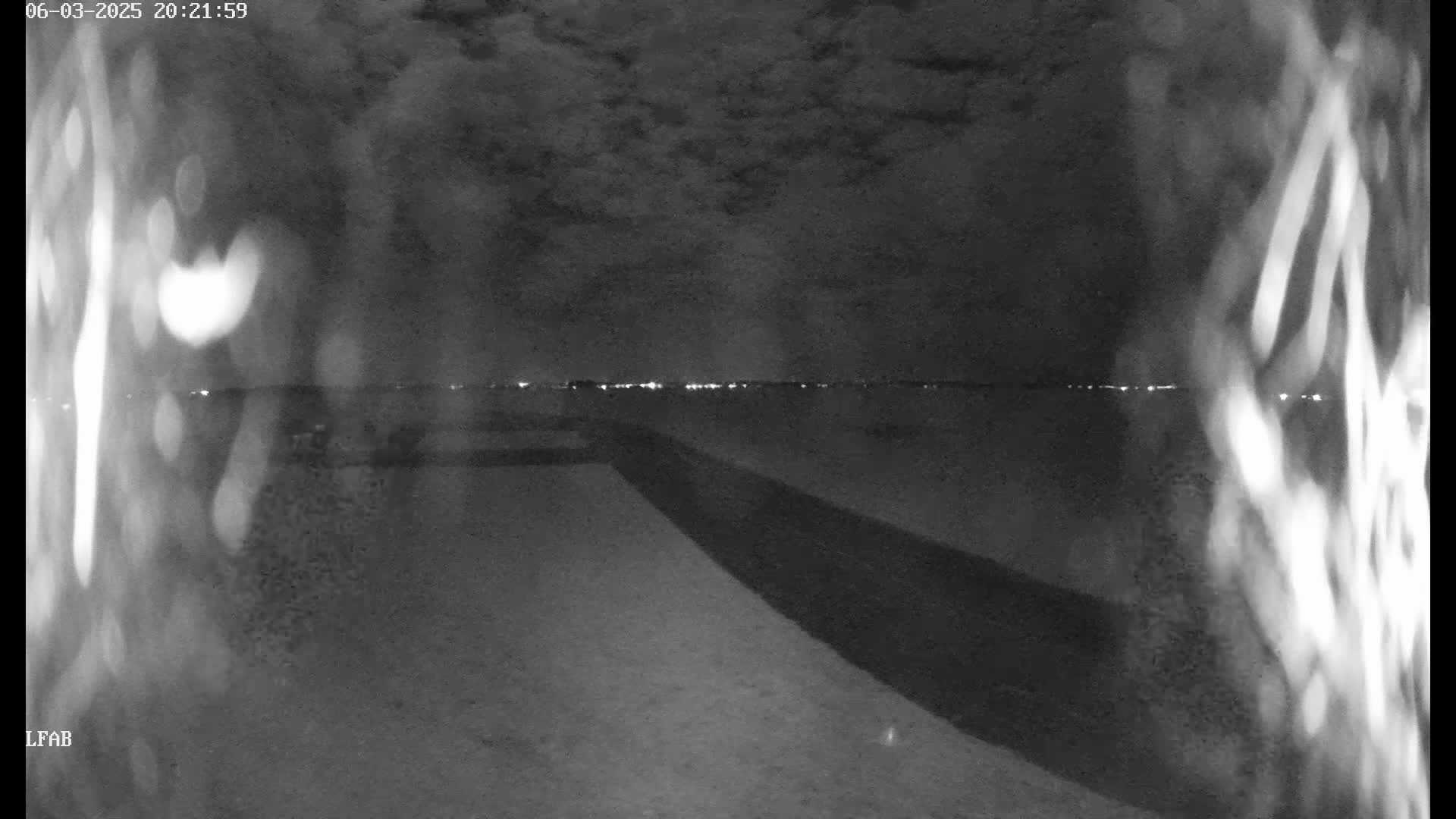 A nighttime, cloudy view shows a dark expanse of land sloping down towards a distant line of lights, possibly a city skyline.