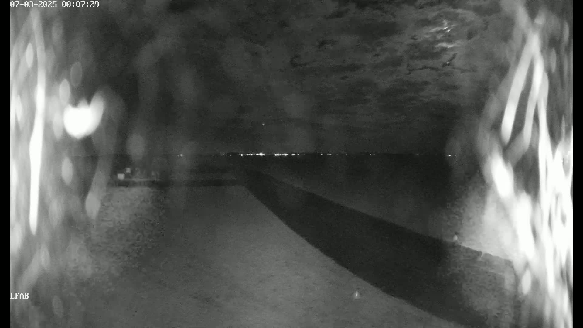 A nighttime, low-light view shows a dark road or path leading toward a distant line of lights under a cloudy sky.
