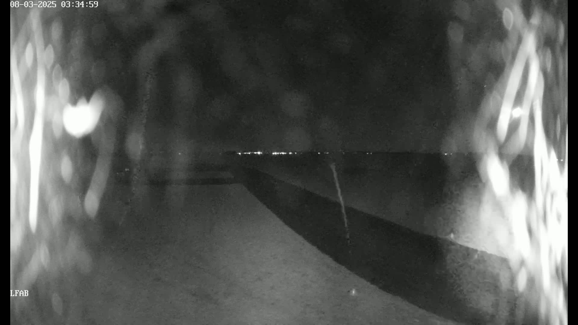 A nighttime, low-light view shows a dark, possibly snowy or icy ground leading to a dark wall or fence, beyond which distant lights are visible.