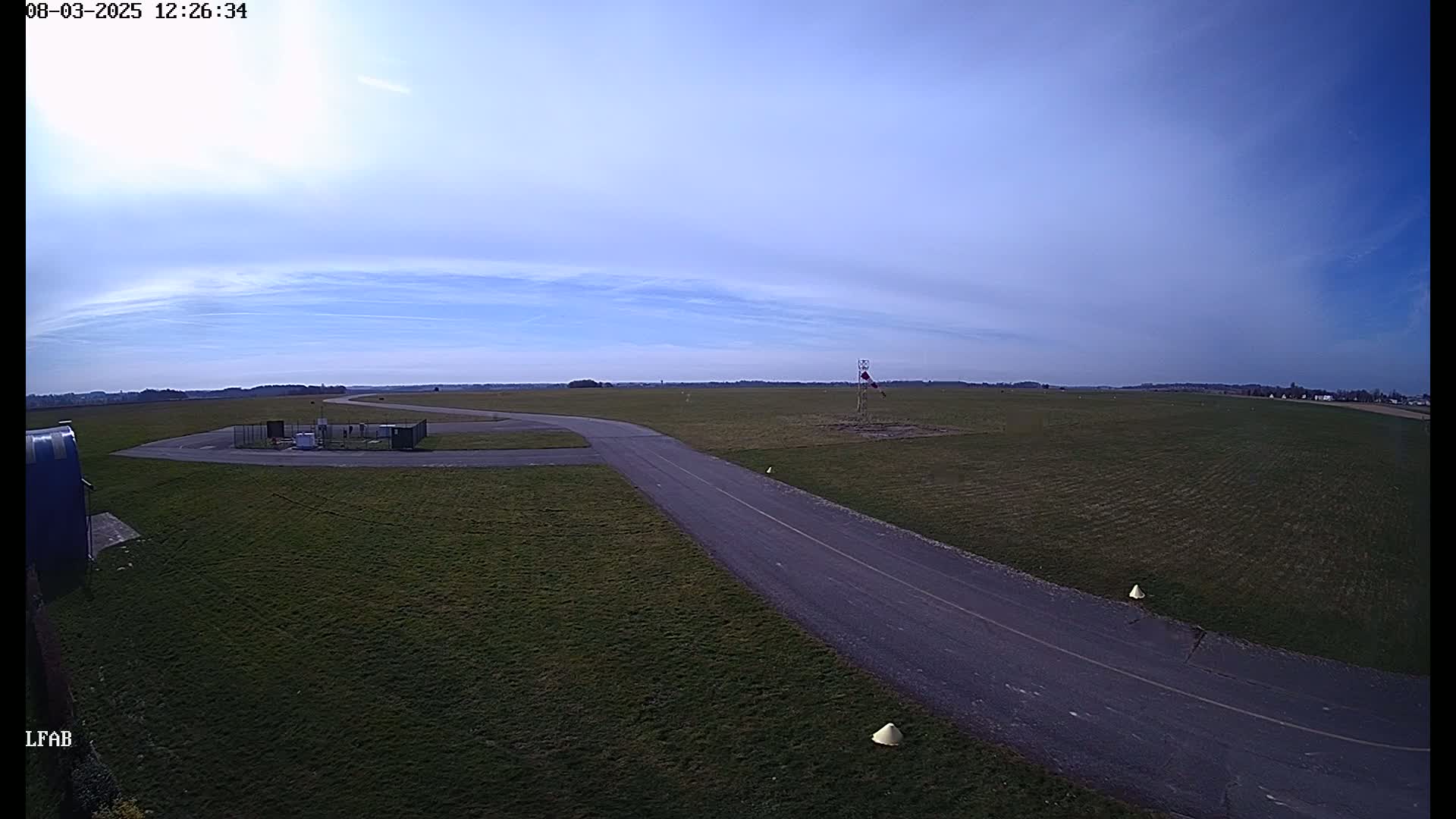 A paved taxiway curves through a grassy airfield under a mostly clear, bright blue sky.