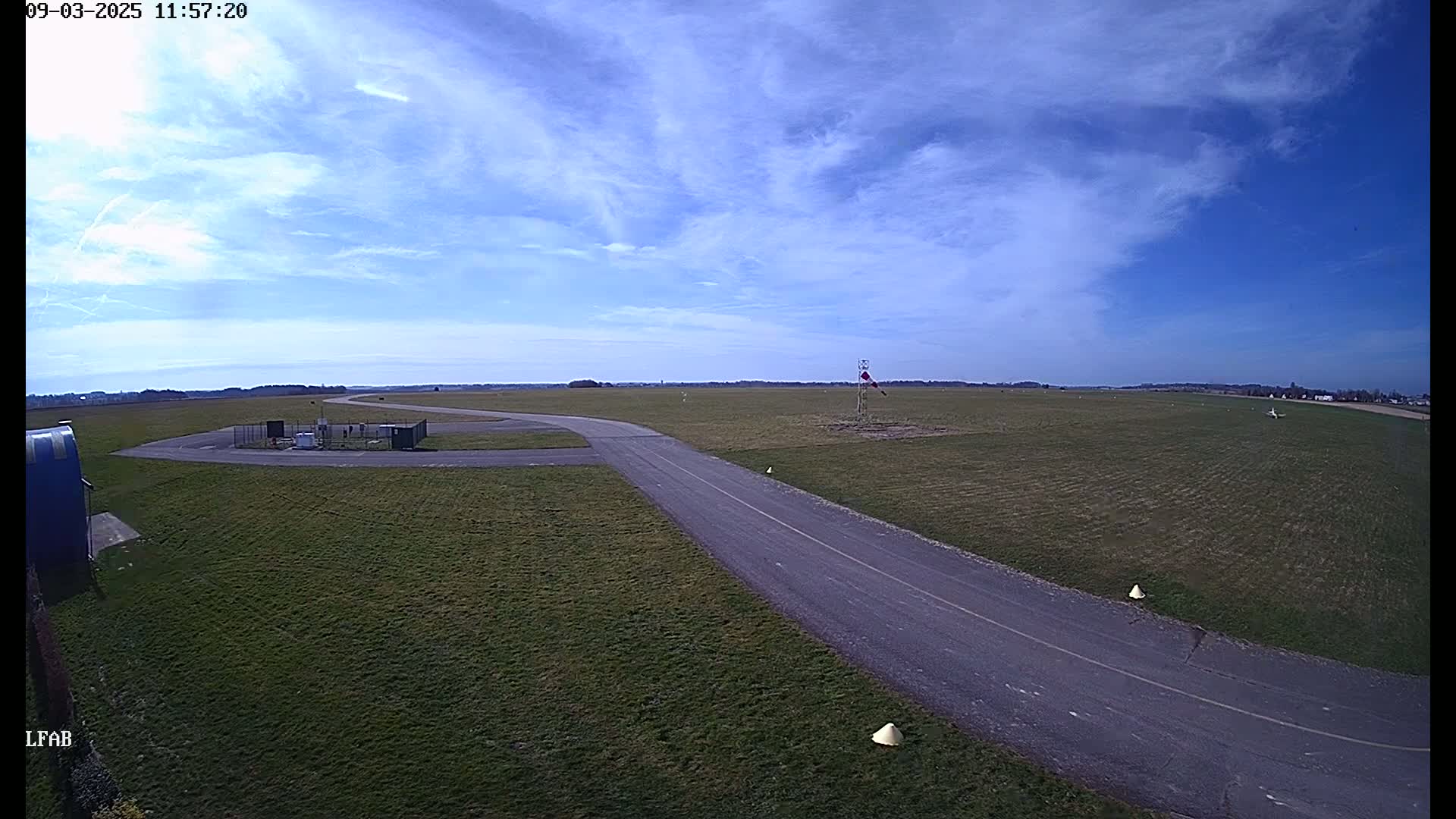 A paved taxiway winds through a grassy airfield under a mostly sunny sky with scattered clouds.