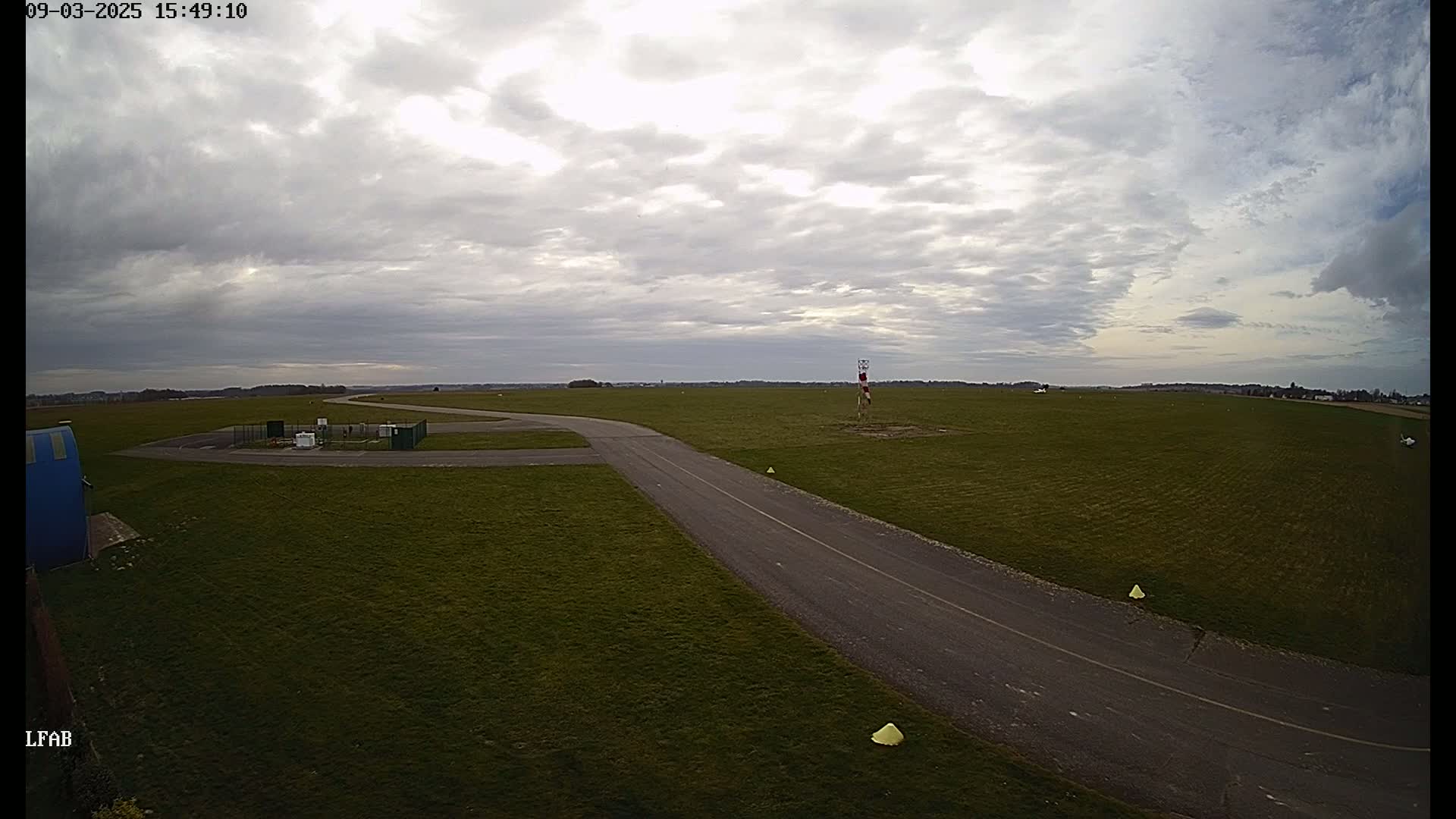An airfield with a paved taxiway is shown under a mostly cloudy sky.