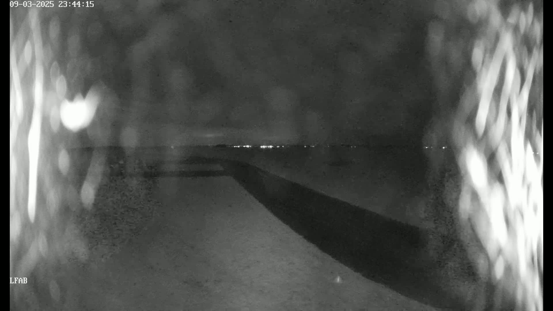 A nighttime view of a dark, flat landscape with a long, dark structure extending into the distance, under a dark, possibly cloudy sky, with faint lights visible on the horizon.