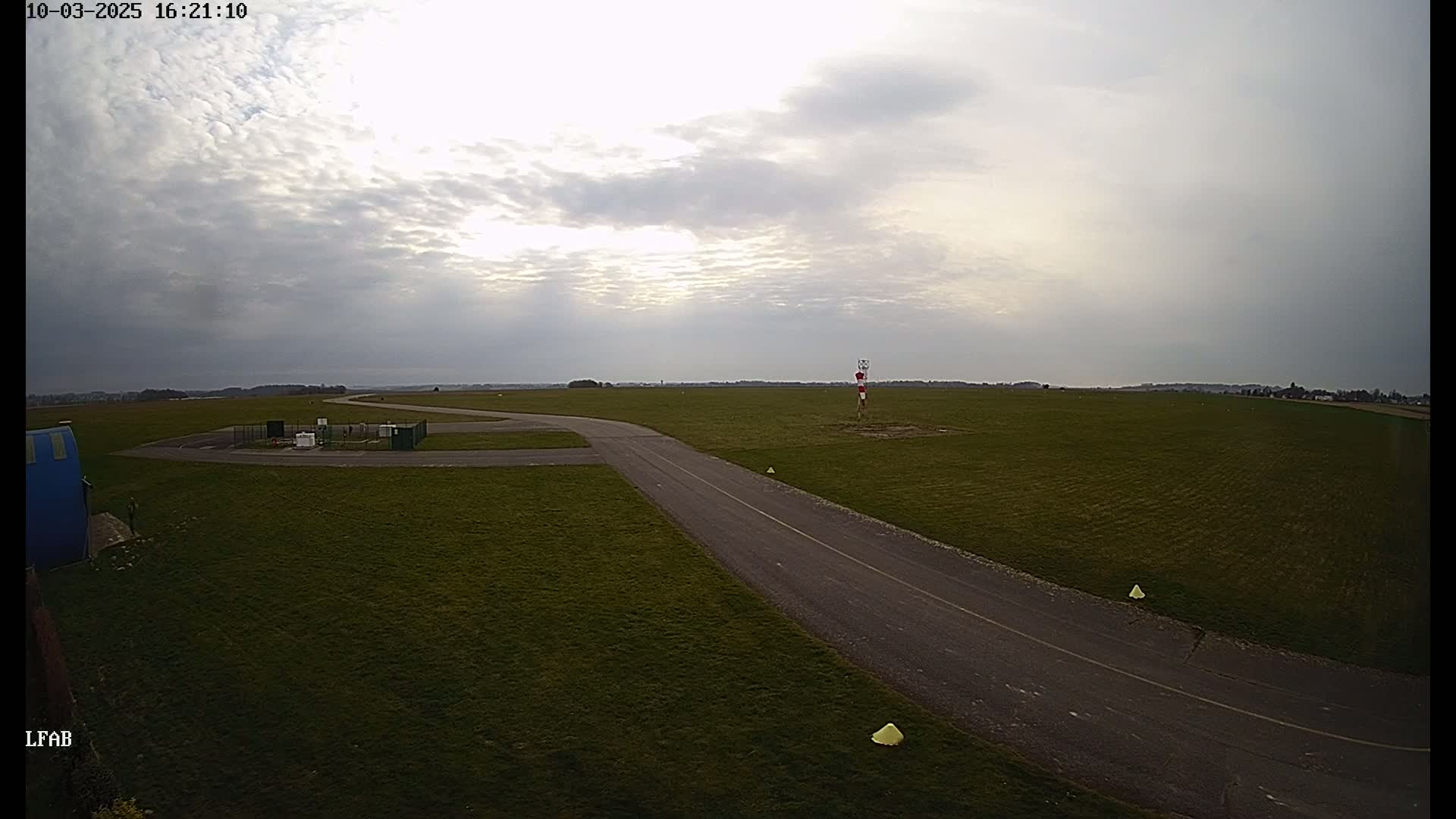 A paved taxiway curves across a grassy airfield under a mostly cloudy sky.