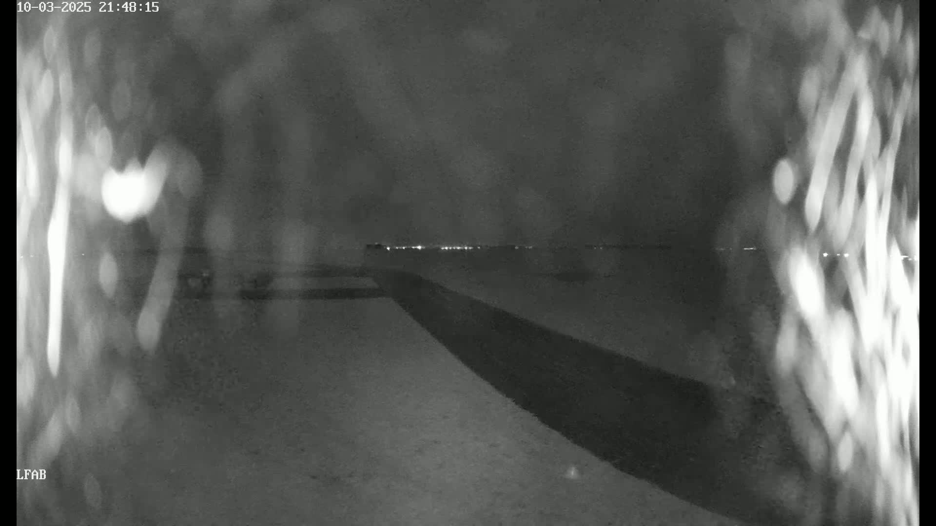 A nighttime, low-light image shows a dark, snow-covered ground with a dark path or road leading towards a distant line of lights.