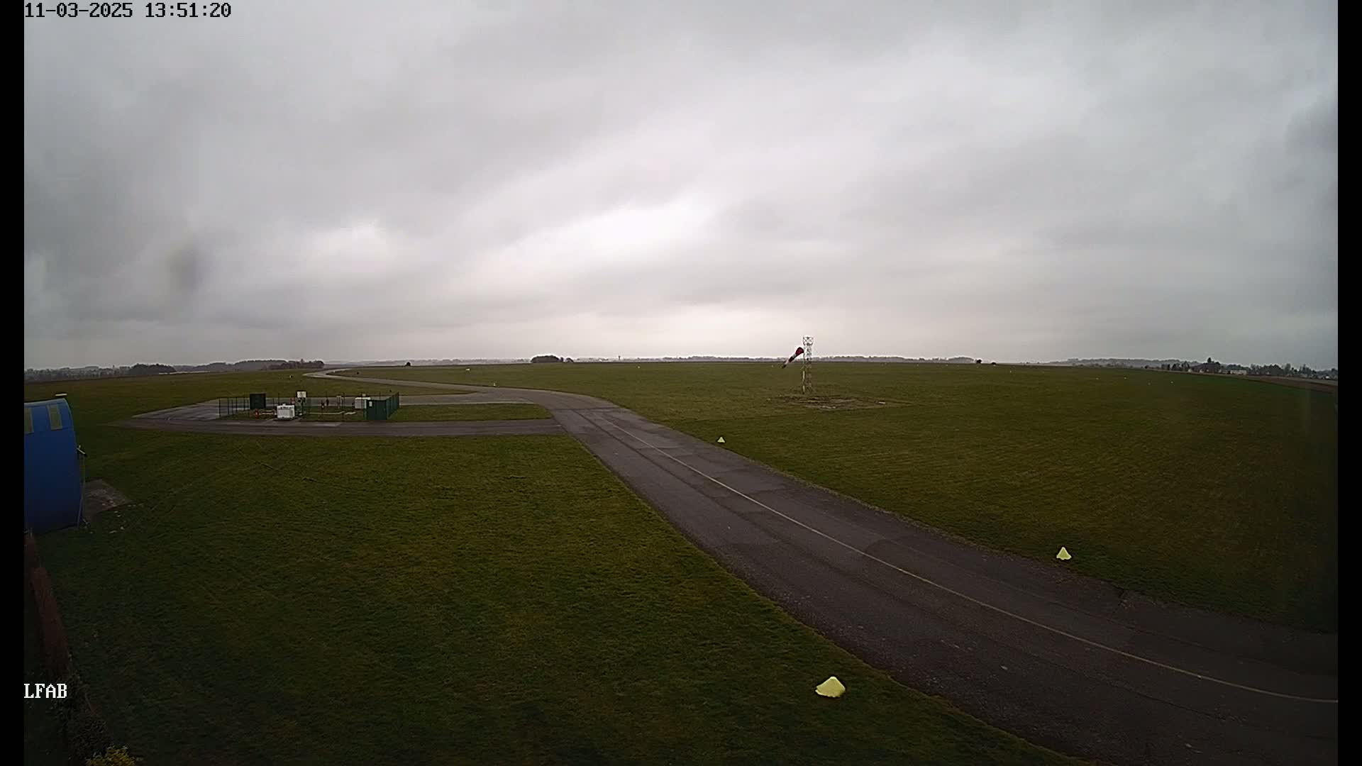 An overcast sky hangs over a grassy airfield with a paved taxiway and windsock.