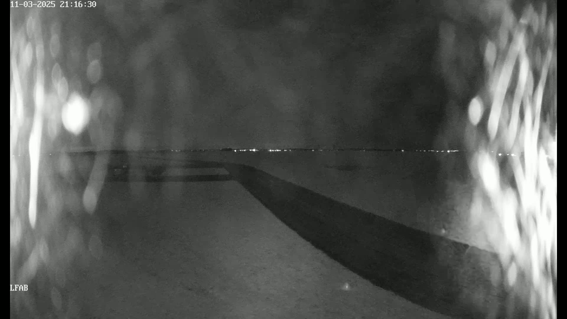A nighttime, low-light view shows a dark, flat expanse of land with a dark road or path stretching towards a distant line of lights on the horizon.