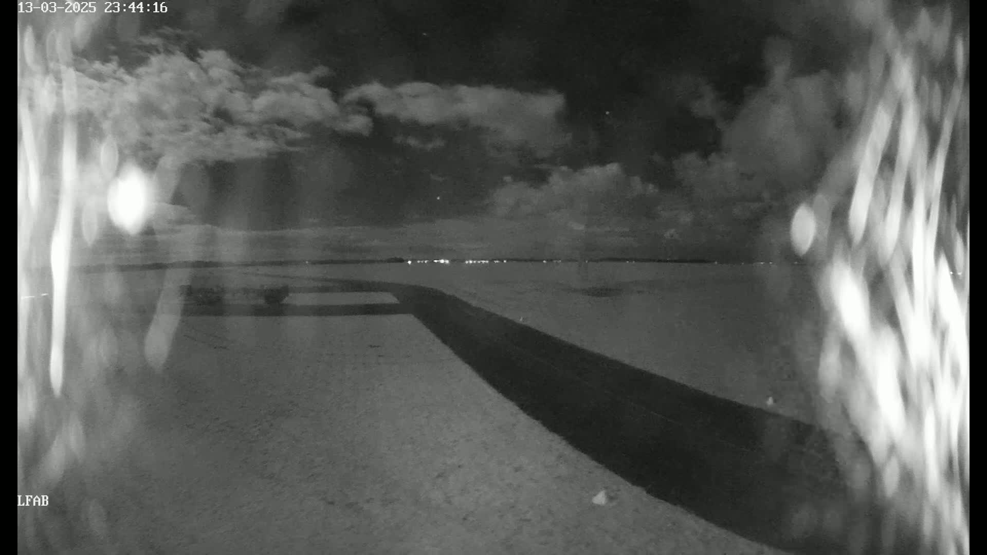 A nighttime, cloudy view shows a dark road stretching toward a distant horizon with faint lights, all set against a flat, lightly textured landscape.