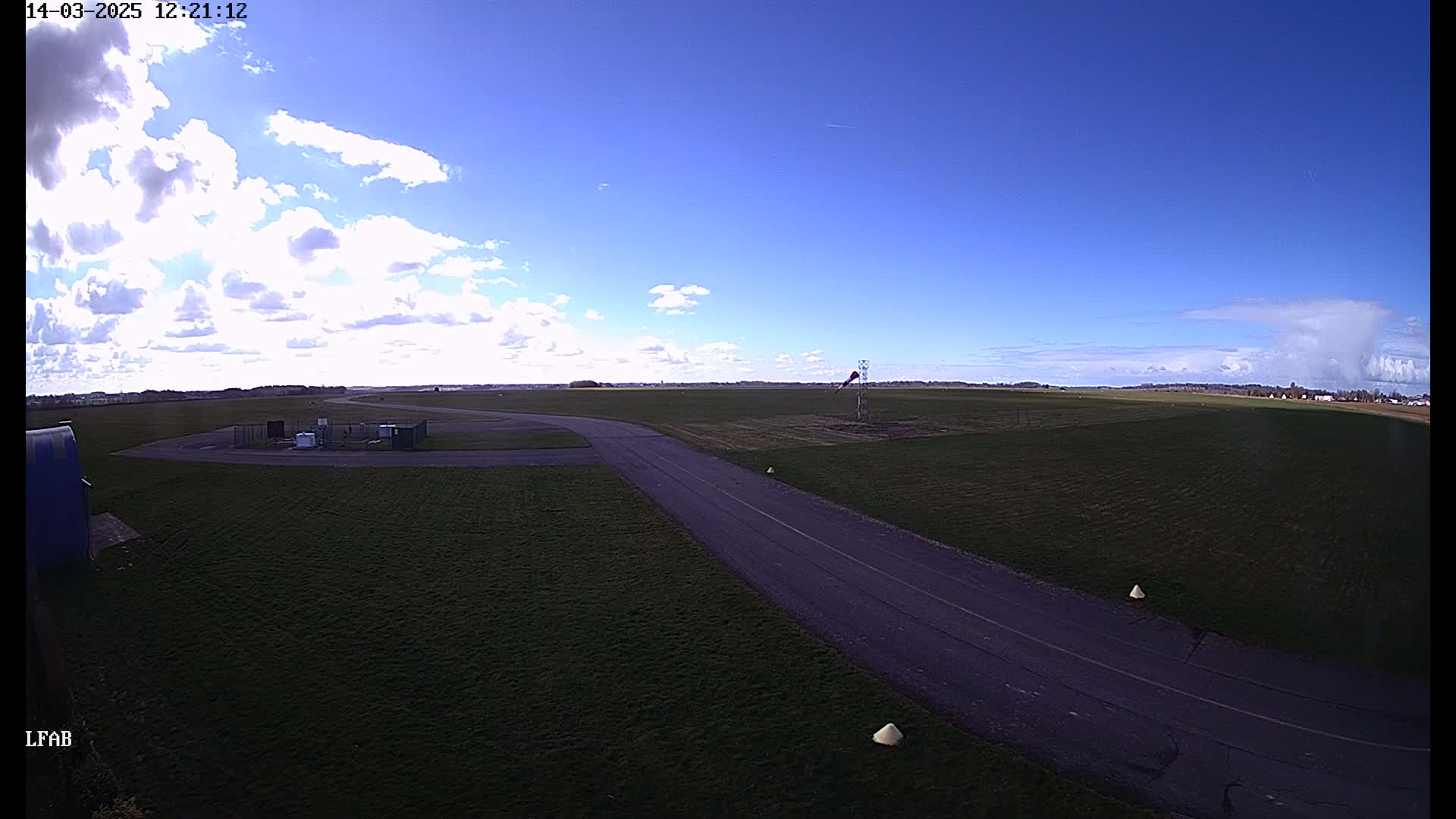 A paved road curves through a grassy airfield under a partly cloudy blue sky.