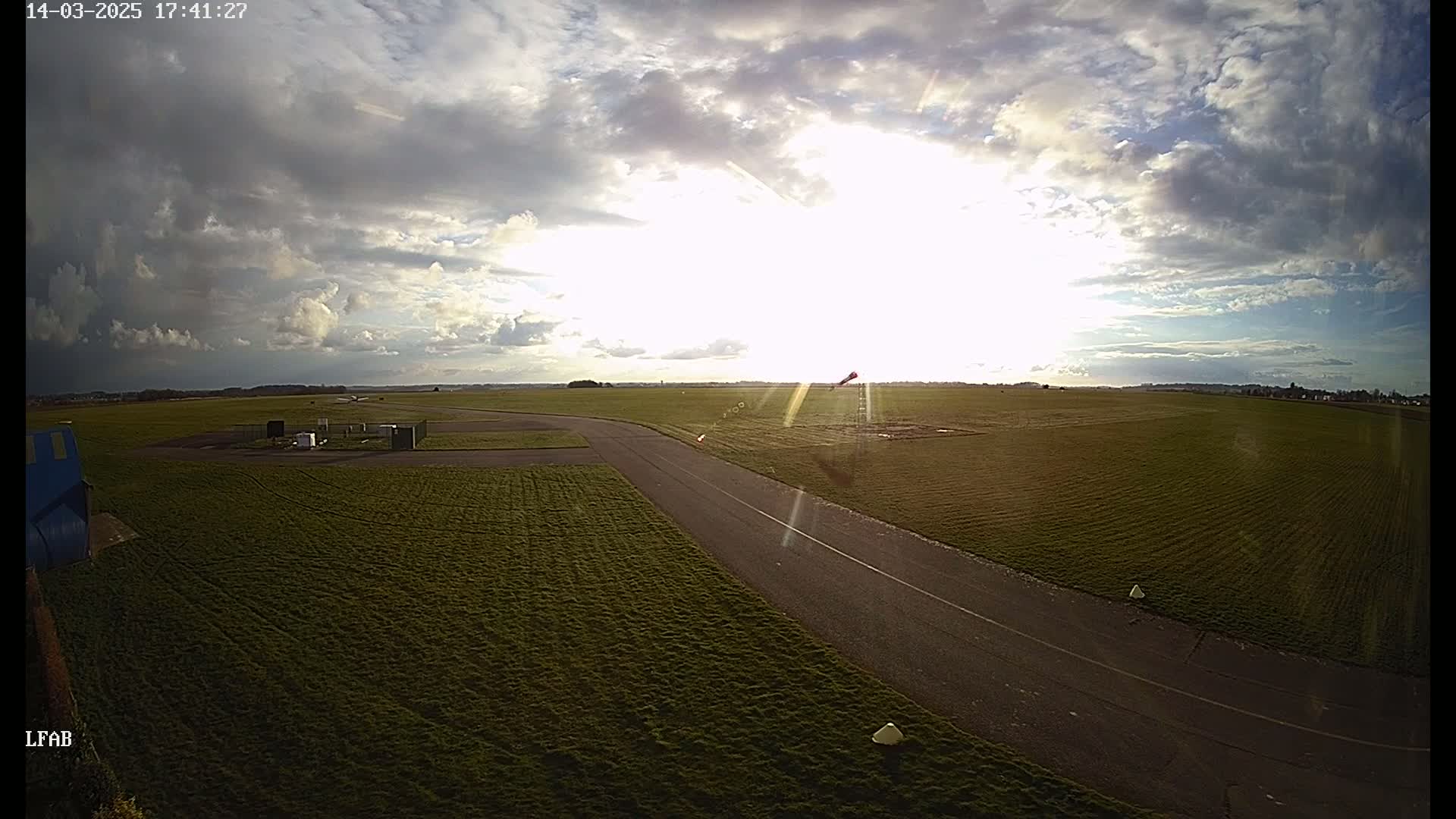 A paved taxiway and grassy airfield are visible under a partly sunny sky with scattered clouds.