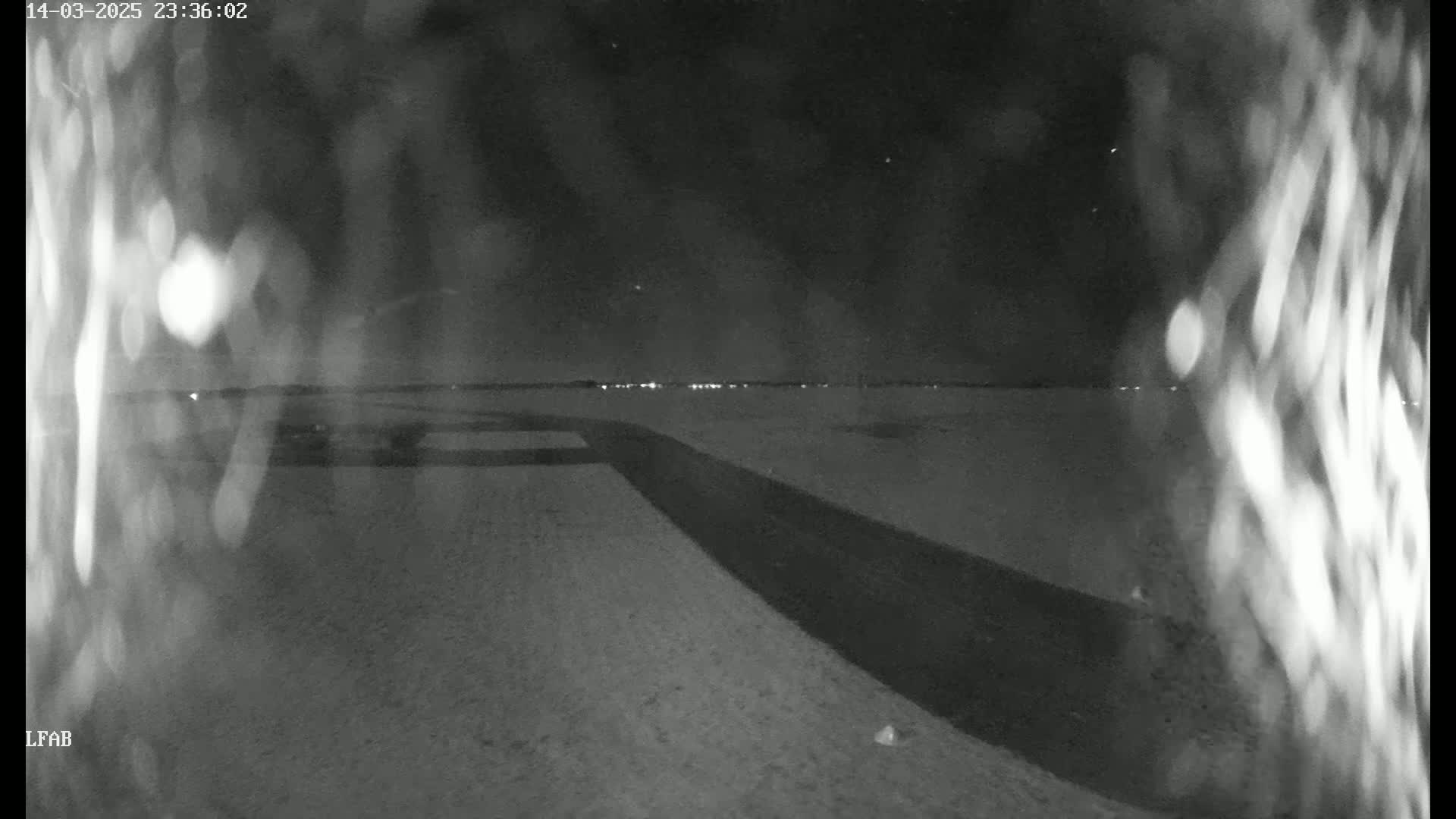 A dark, snow-covered area at night, with a dark road or path leading towards a distant horizon faintly lit by lights.