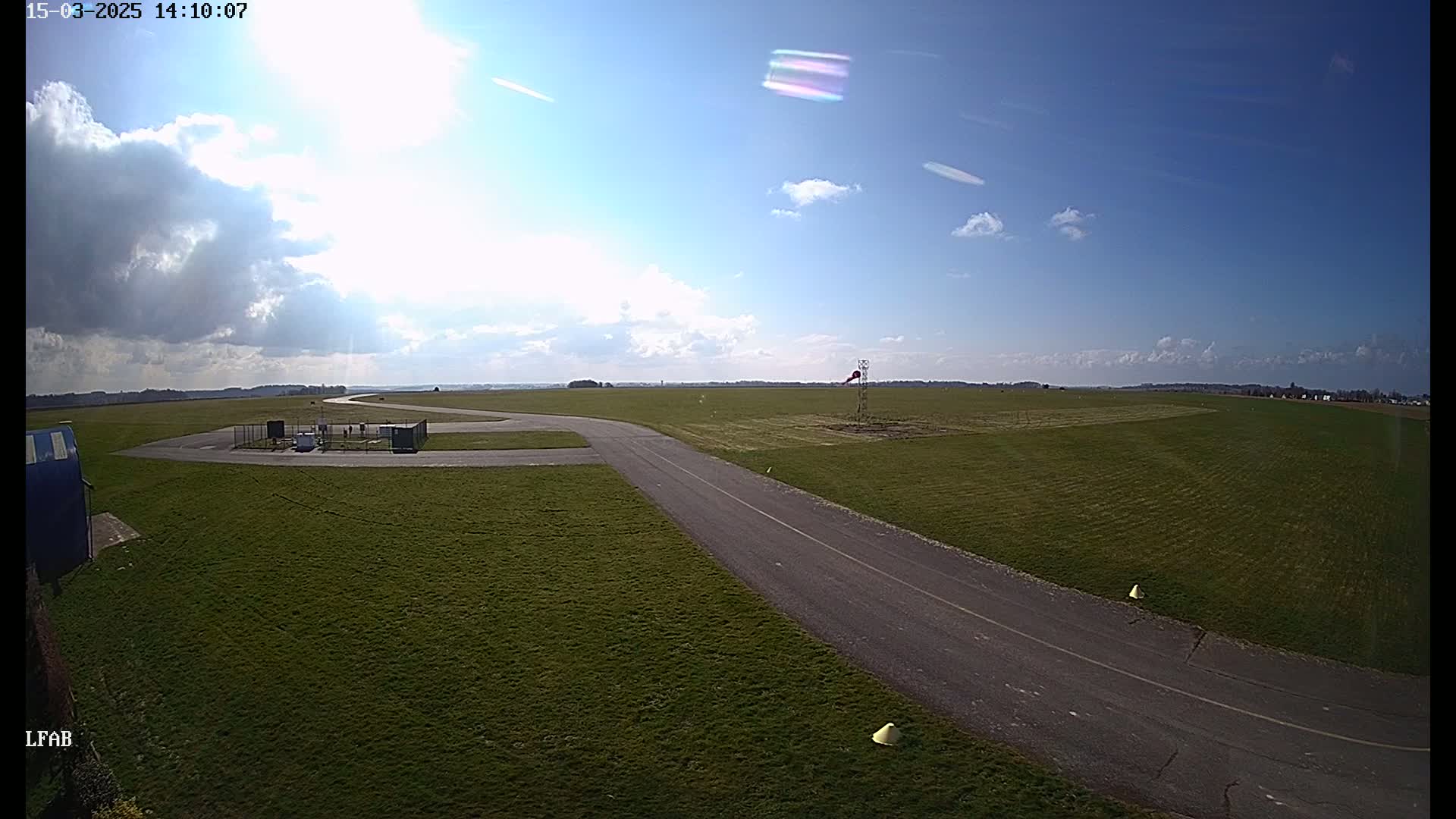 A paved taxiway curves through a grassy airfield under a mostly sunny sky with scattered clouds.