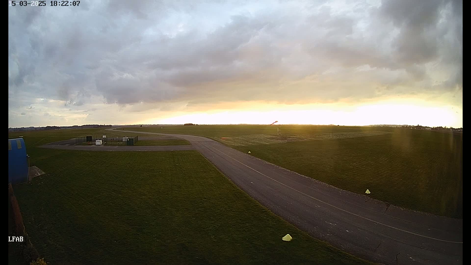 A paved taxiway curves through a grassy airfield under a partly cloudy sky with a setting sun.