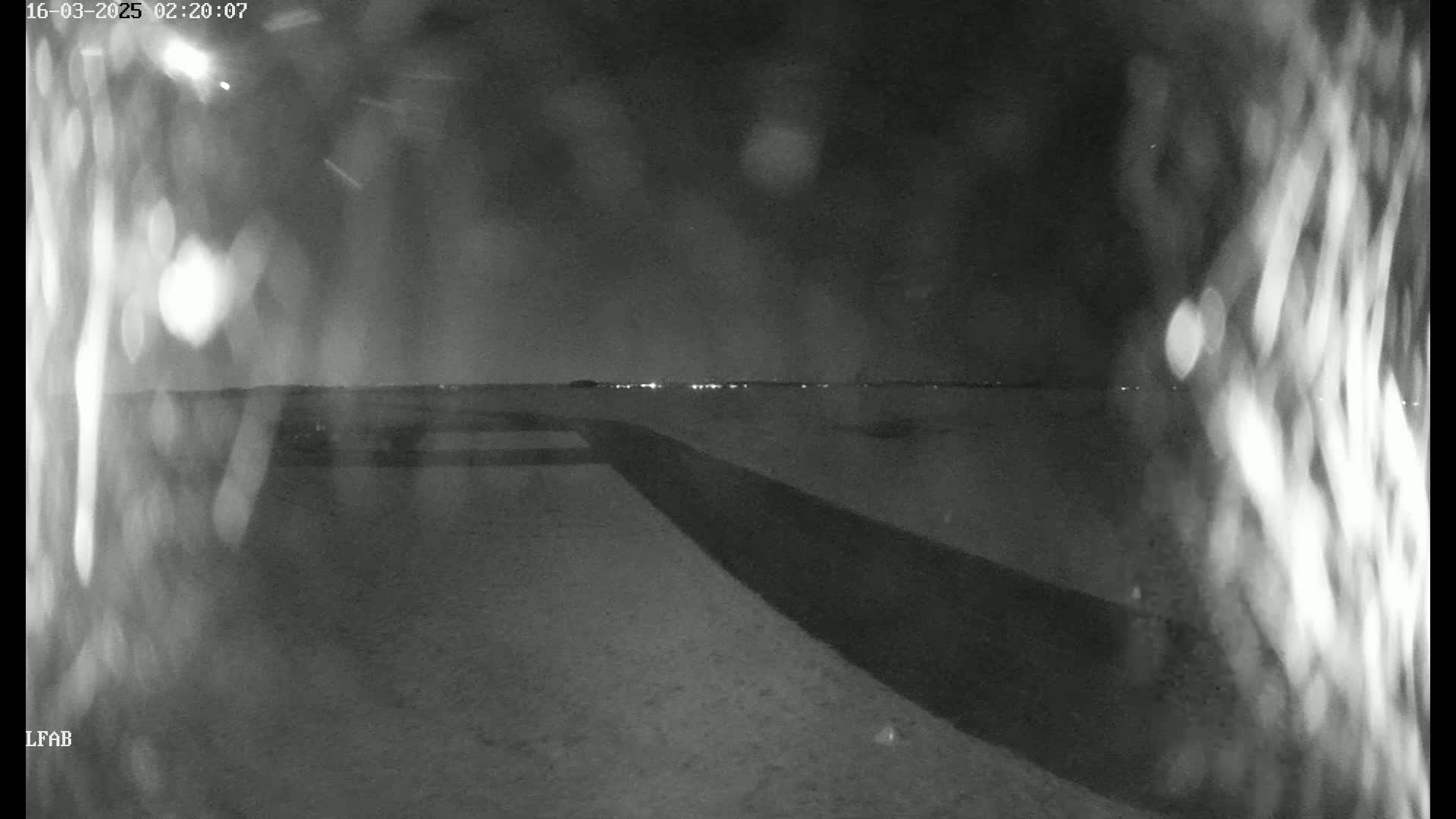 A nighttime, low-light view shows a dark, flat expanse of land with a dark road or path curving into the distance, under a dark sky with faint lights visible on the horizon.