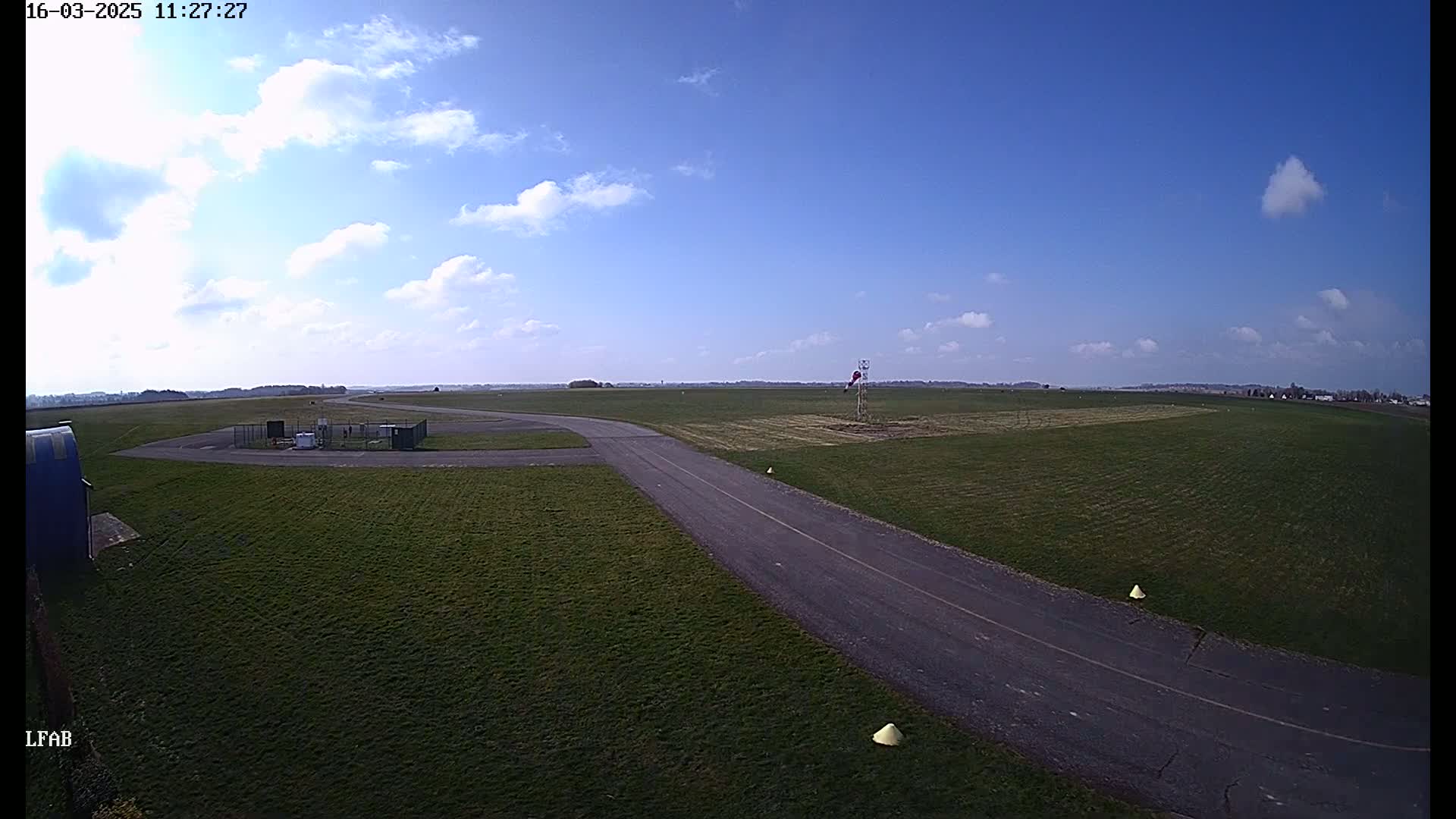 A paved taxiway curves through a grassy airfield under a mostly clear blue sky with scattered cumulus clouds.