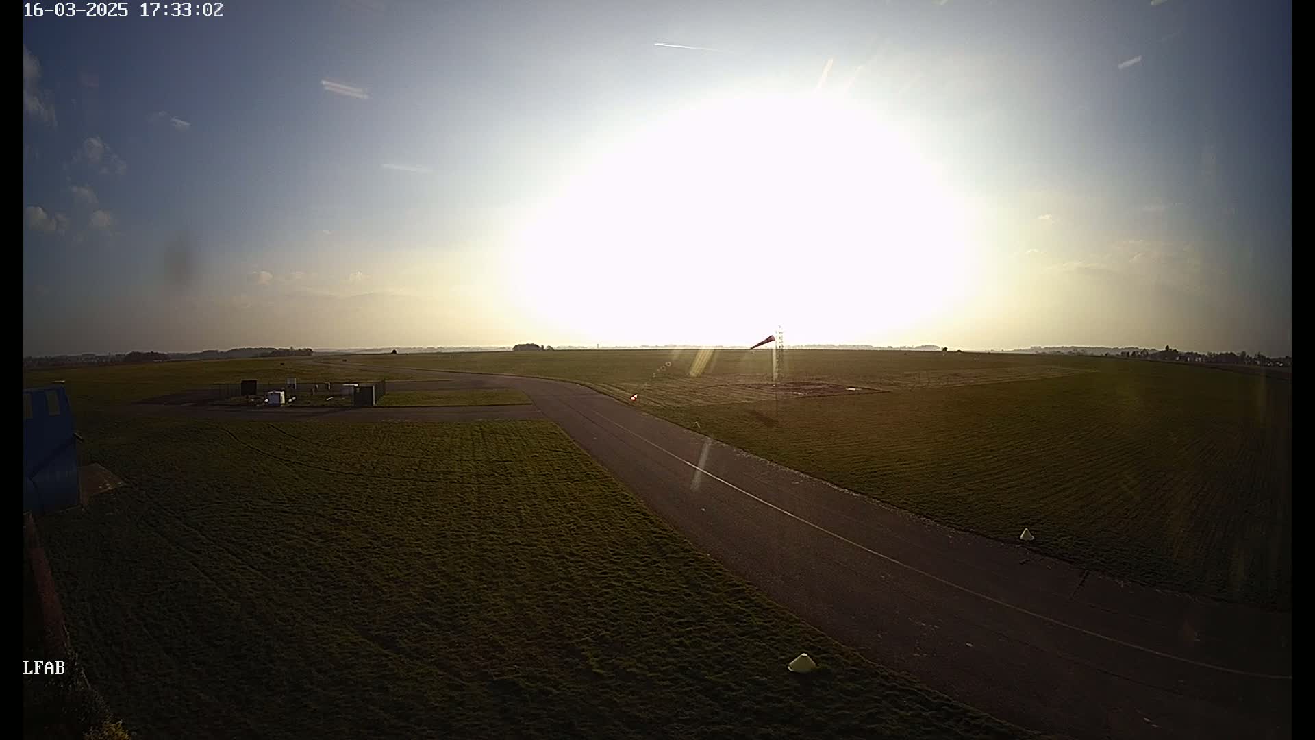 A paved roadway curves through a grassy airfield under a bright, sunny sky with a few wispy clouds.