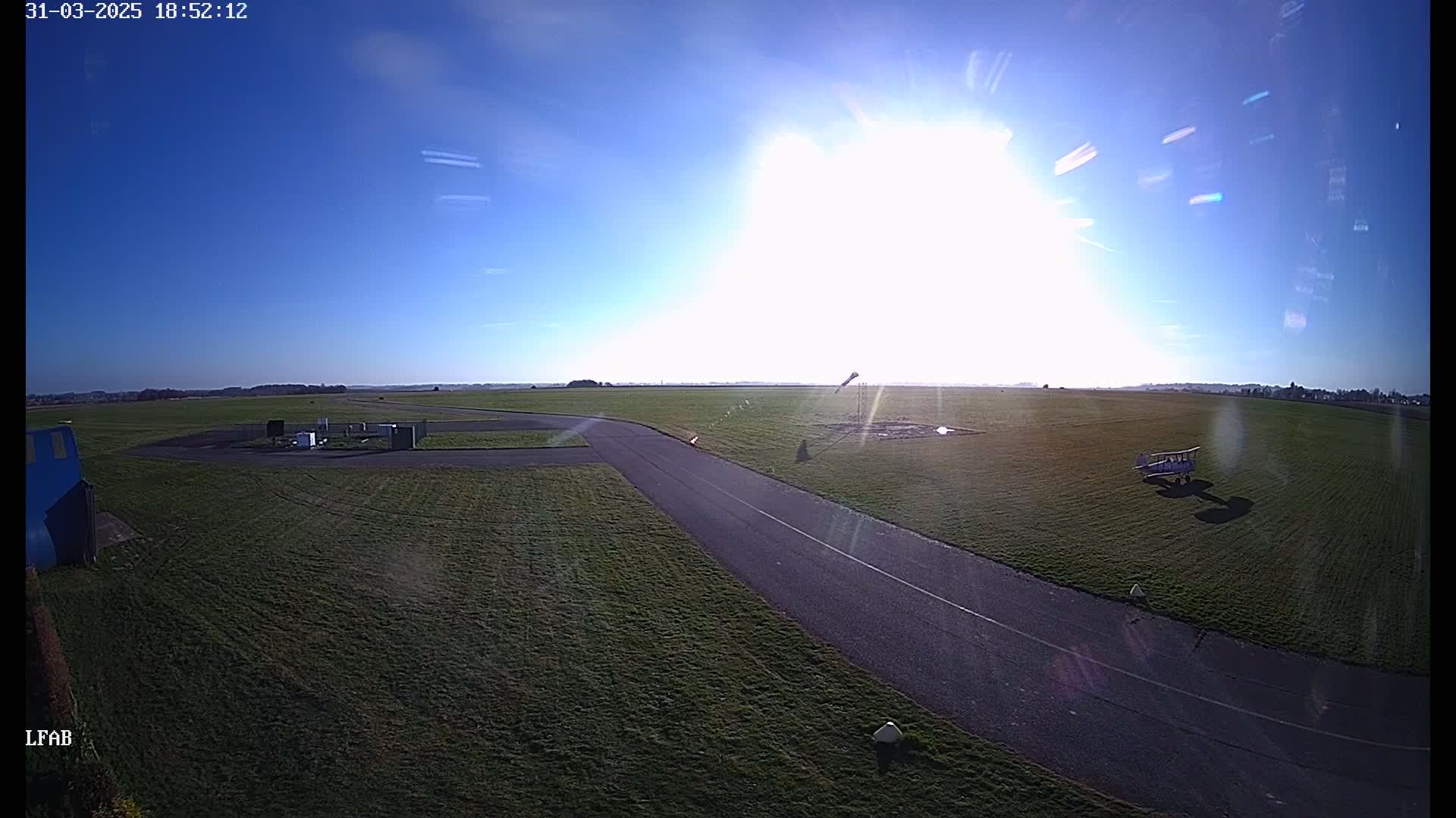 A bright sunny day reveals a grassy airfield with a small biplane parked on the grass near a paved taxiway.