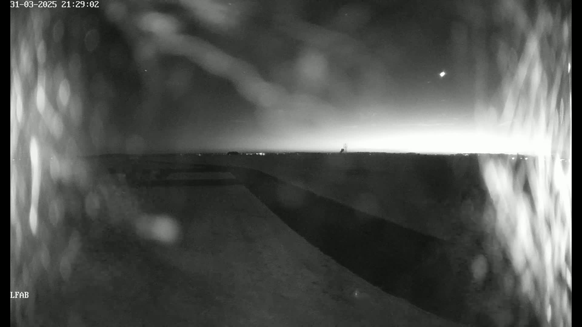 A nighttime view of a flat, dark landscape under a mostly clear sky, with rain or moisture obscuring the foreground.
