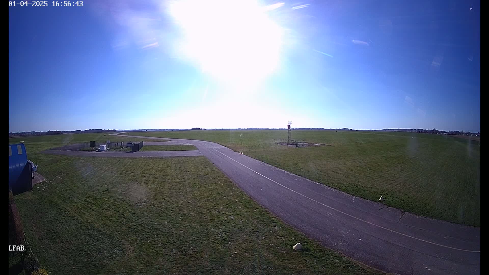 A sunny day reveals a paved taxiway curving through a grassy airfield, with small buildings and equipment visible nearby.