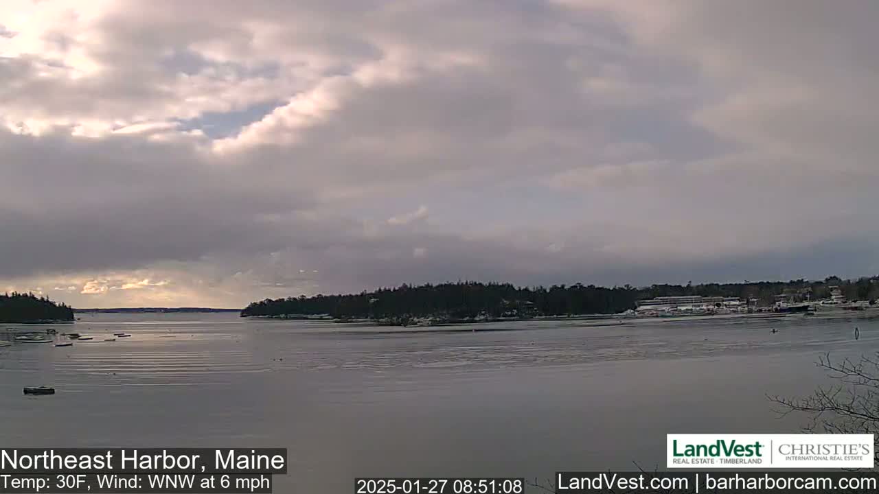 A partly cloudy, cold day with a light wind over a partially frozen harbor, with snow visible on the far shore and a few boats scattered in the water.
