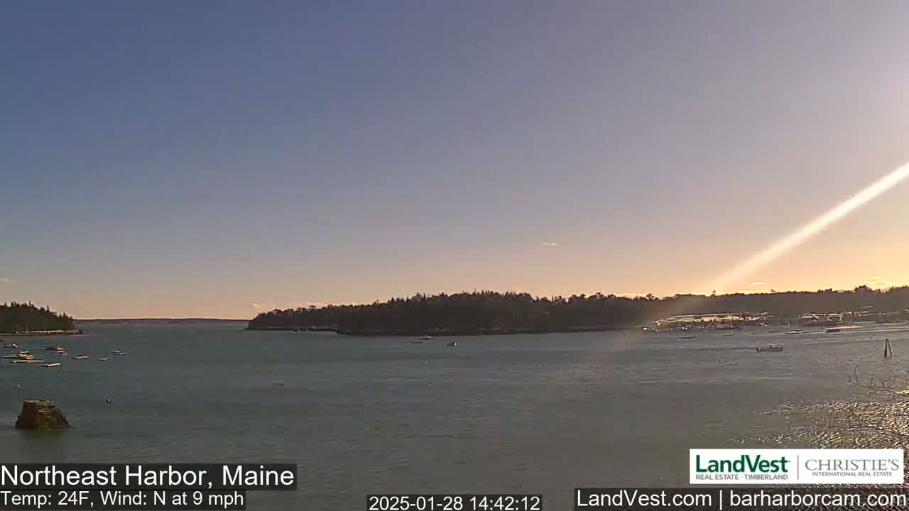A calm harbor with a few boats is viewed at sunset under a clear, pale blue sky with a sunbeam extending across the water.