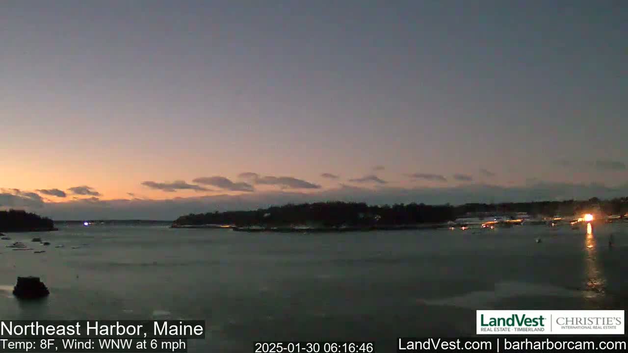 A calm body of water at dusk, under a mostly clear, twilight sky, with a small island in the distance and some lights visible along the shoreline.