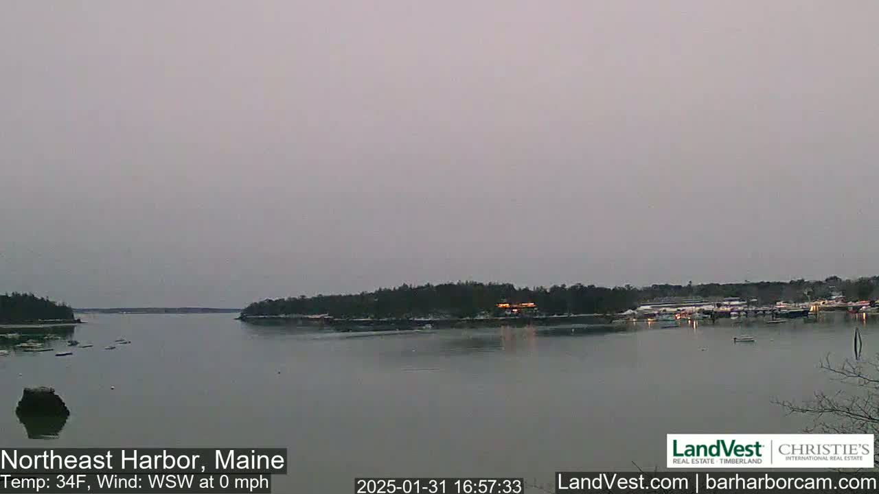 A calm, overcast harbor scene shows a body of water with snow-dusted land and a few boats.