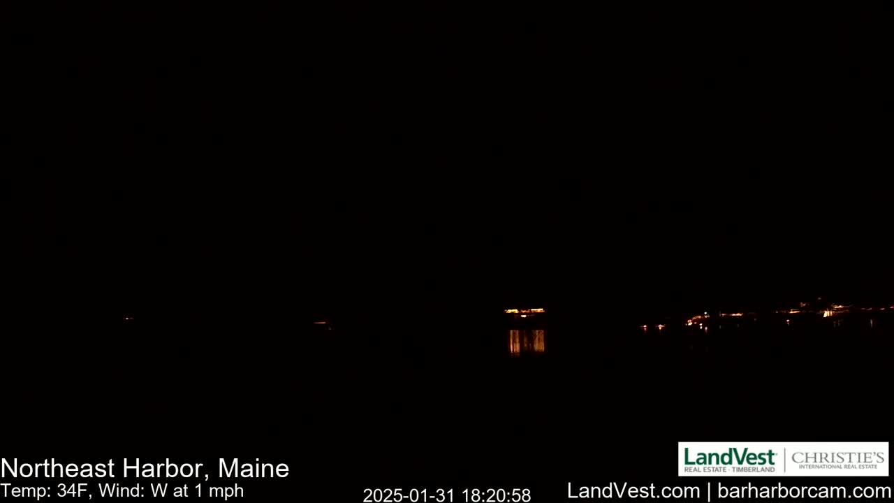 A dark nighttime scene shows a few distant lights across a body of water under a clear, dark sky with a light west wind.