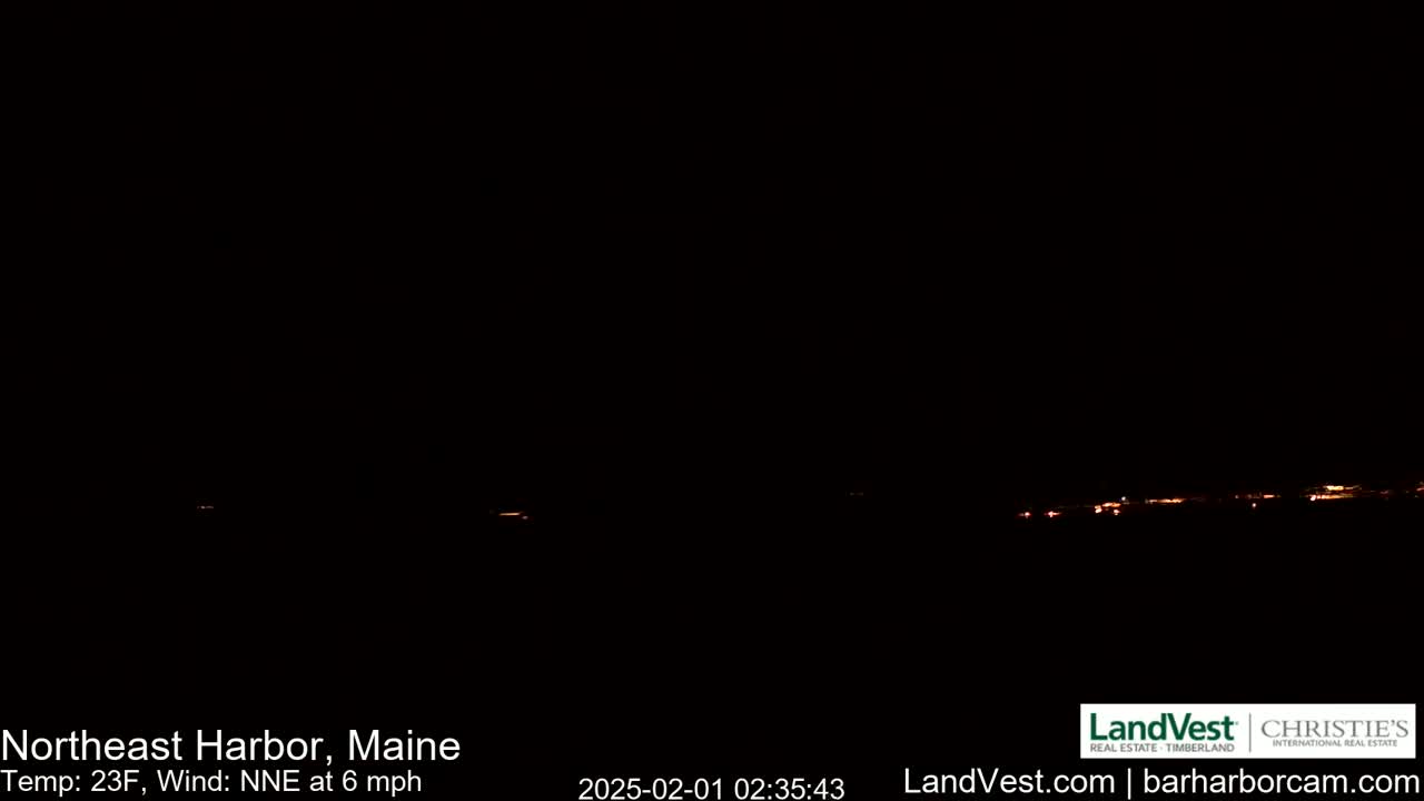 A nighttime long shot shows a distant line of faint orange lights across a dark horizon under a clear, dark sky, with a temperature of 23°F and a north-northeast wind at 6 mph.