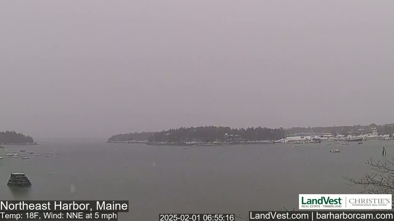A snow-covered harbor scene shows a calm body of water with a few boats and a snow-dusted shoreline under an overcast sky.