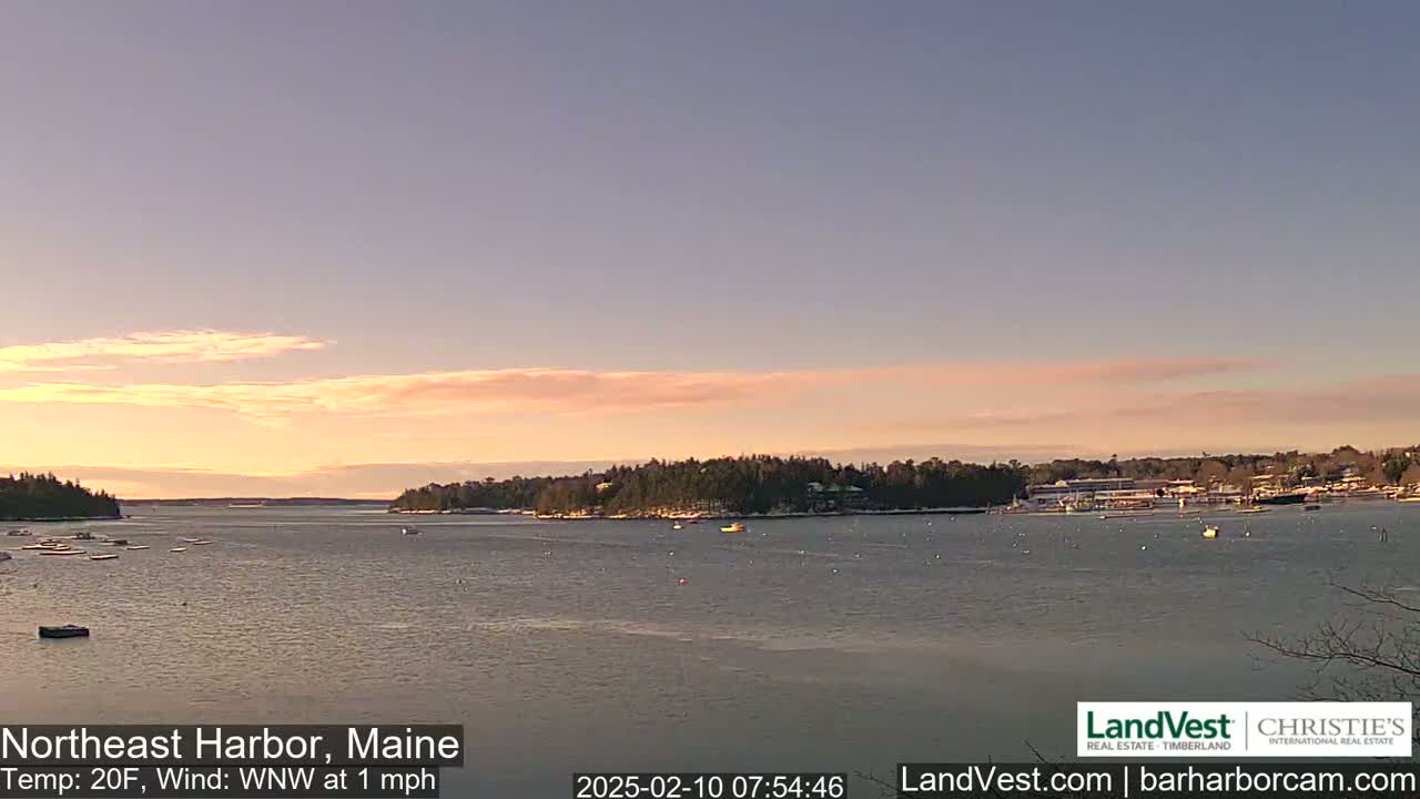 A calm harbor with a few boats is seen at sunrise under a pale pink and orange sky, with a light wind.