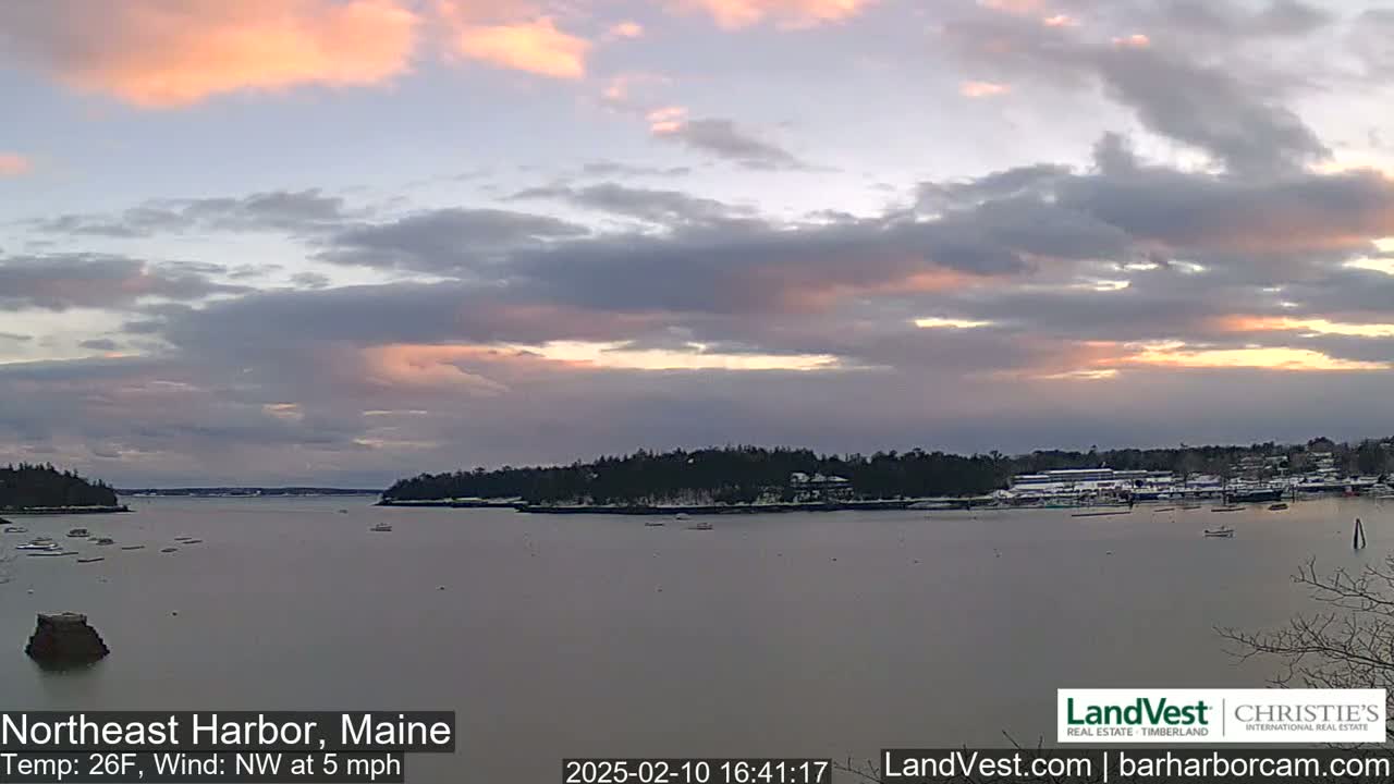 A calm body of water is seen at sunset under a partly cloudy sky, with snow-covered trees and buildings visible in the distance.
